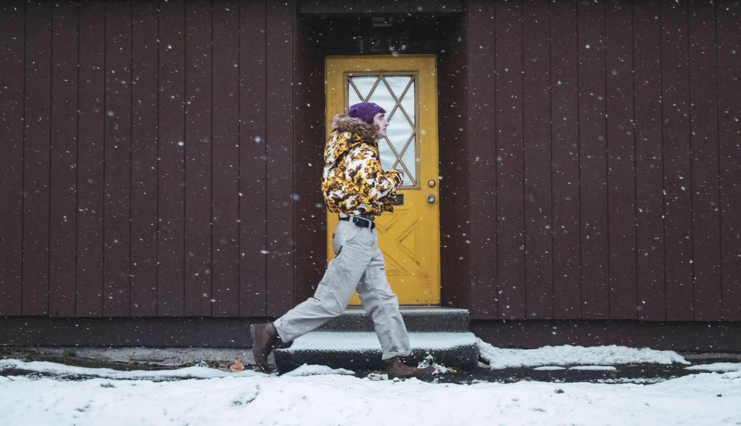 Person in floral yellow jacket and purple beanie walks past yellow door in snow.