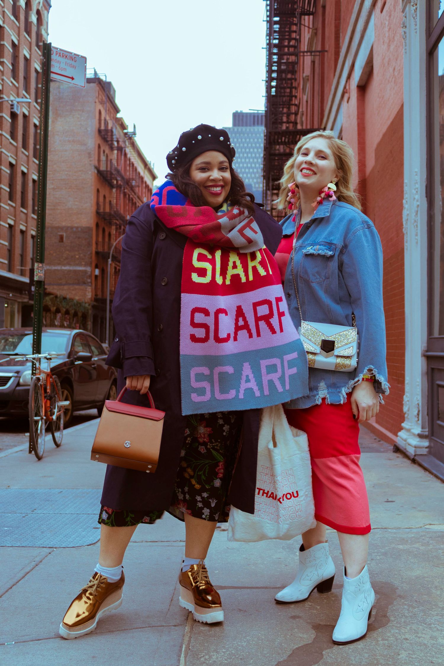 Two stylish women pose on city street, one wearing colorful SCARF-printed wrap accessory.