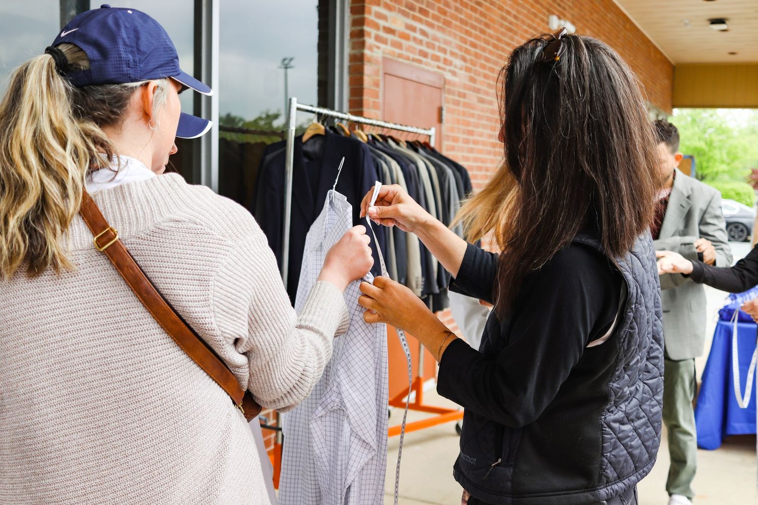 Two women browse shirts on a clothing rack outside a brick building at a community event.