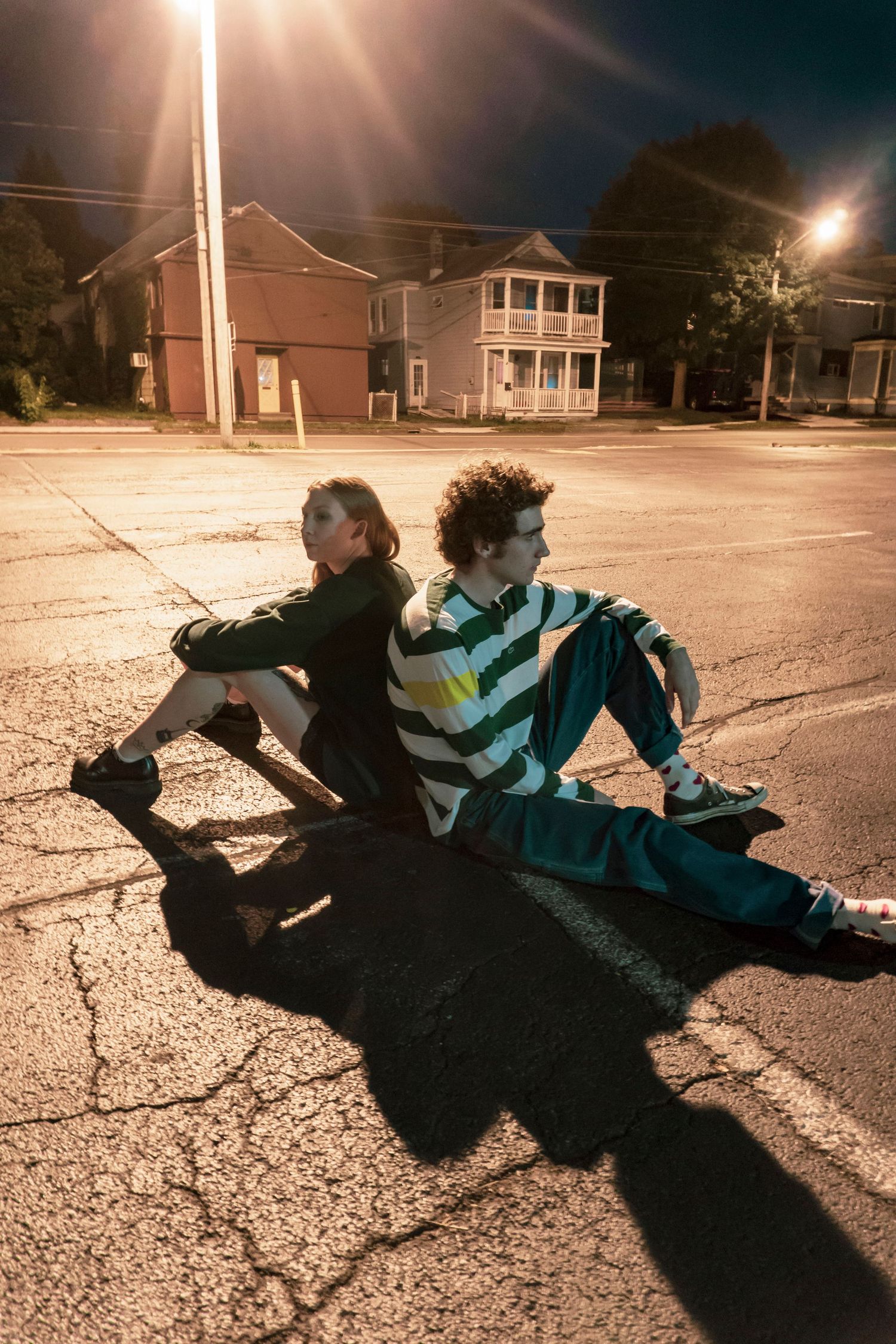 Two people sit back-to-back on a cracked street at night under bright streetlights.