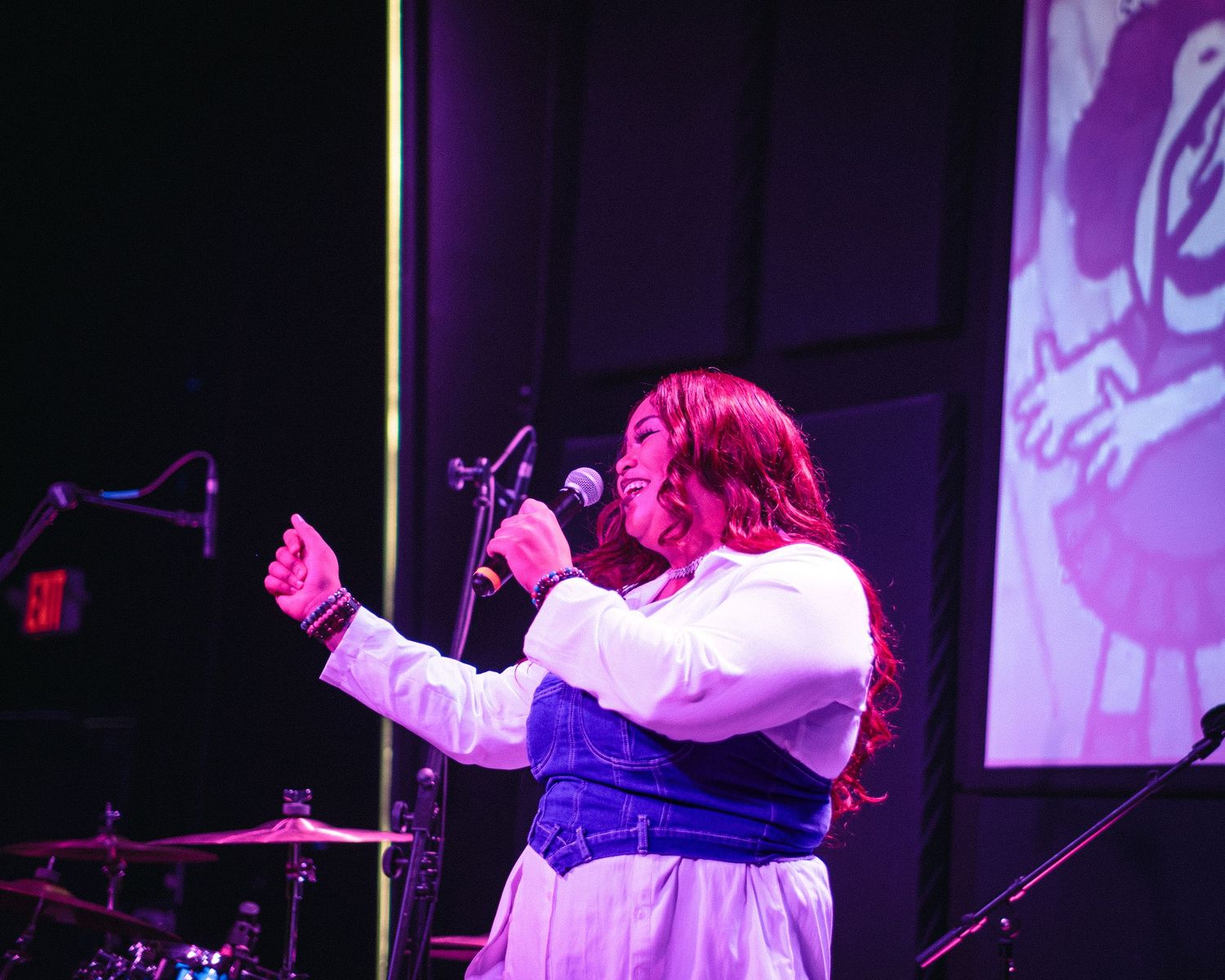 A female vocalist sings passionately into a microphone on a purple-lit stage.