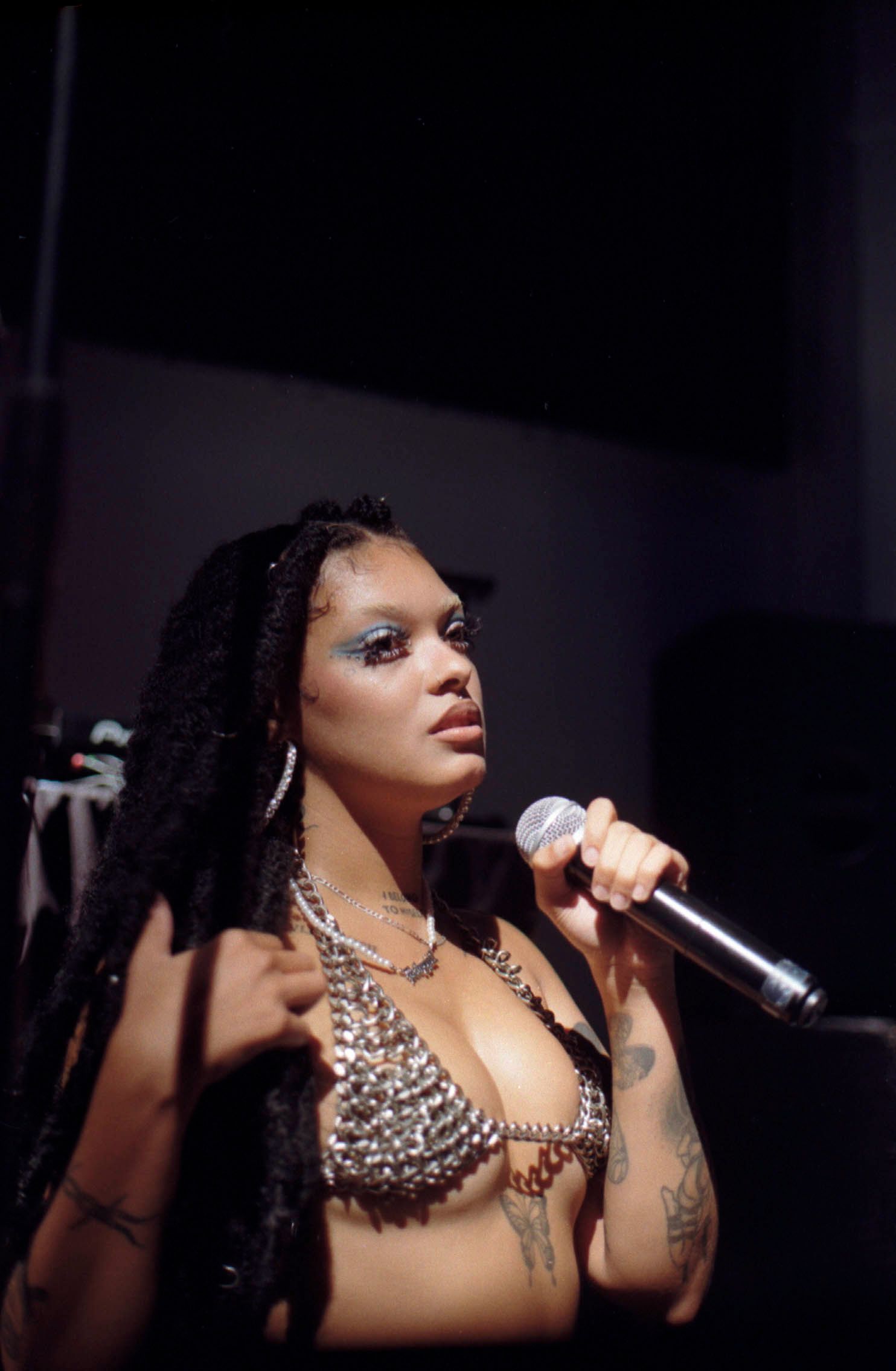 Female performer singing into microphone wearing rhinestone bra with locs and dramatic eye makeup.