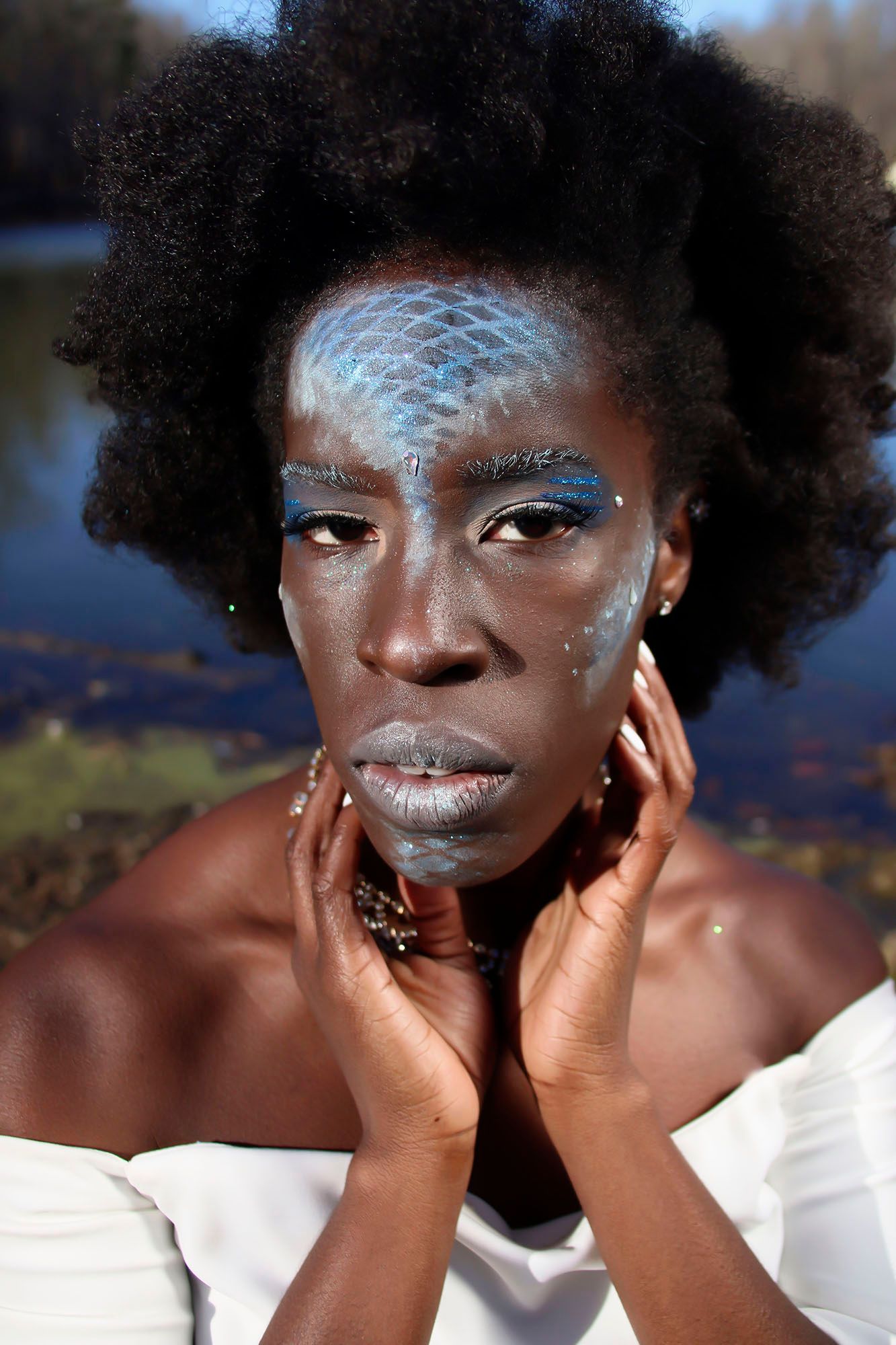 A dark-skinned woman with blue scale-patterned face paint and silver lips poses outdoors in white top.