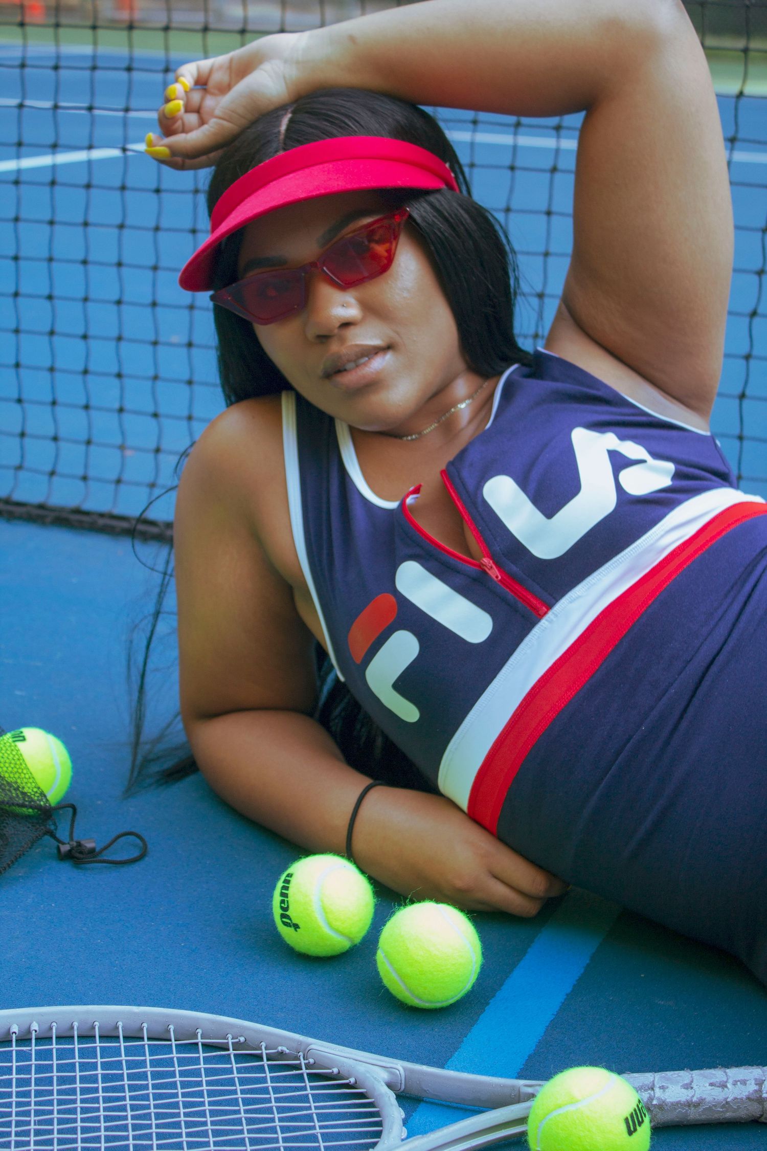 Woman in FILA tennis dress and red visor posing on blue court surrounded by tennis balls.