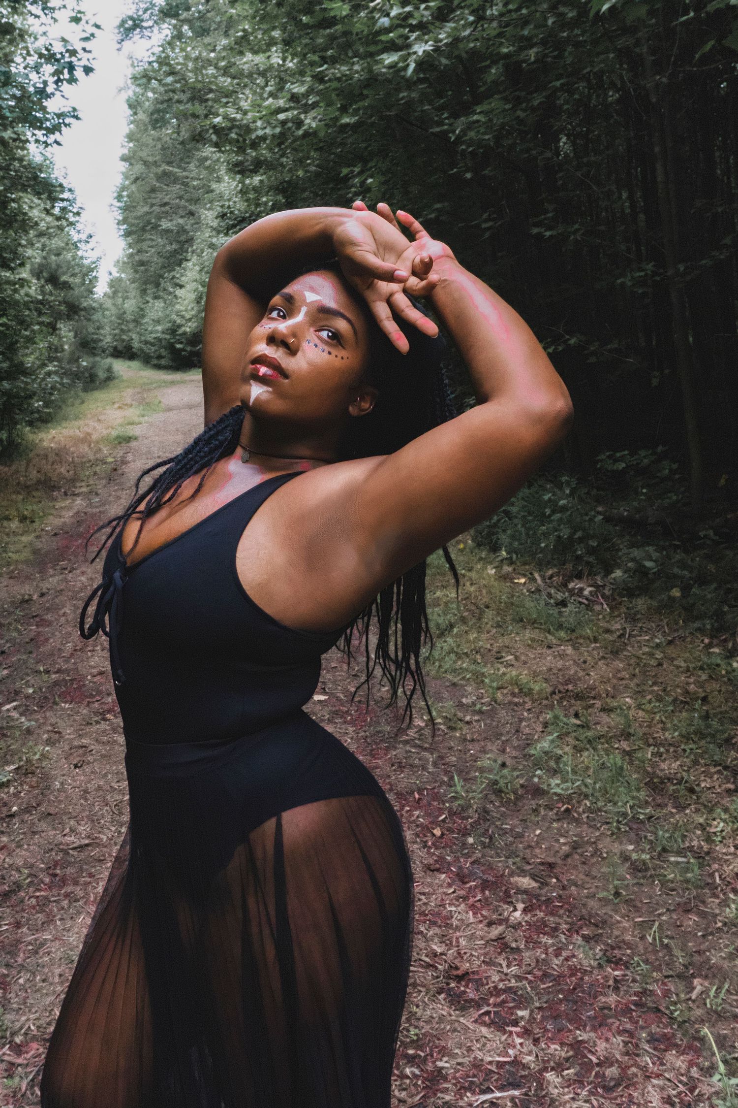 Black woman in sheer black dress with face paint poses dramatically on forest trail.