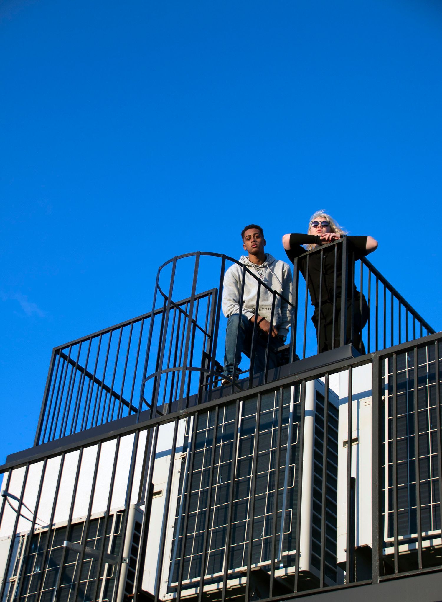 Two people standing on a rooftop fire escape against a clear blue sky.