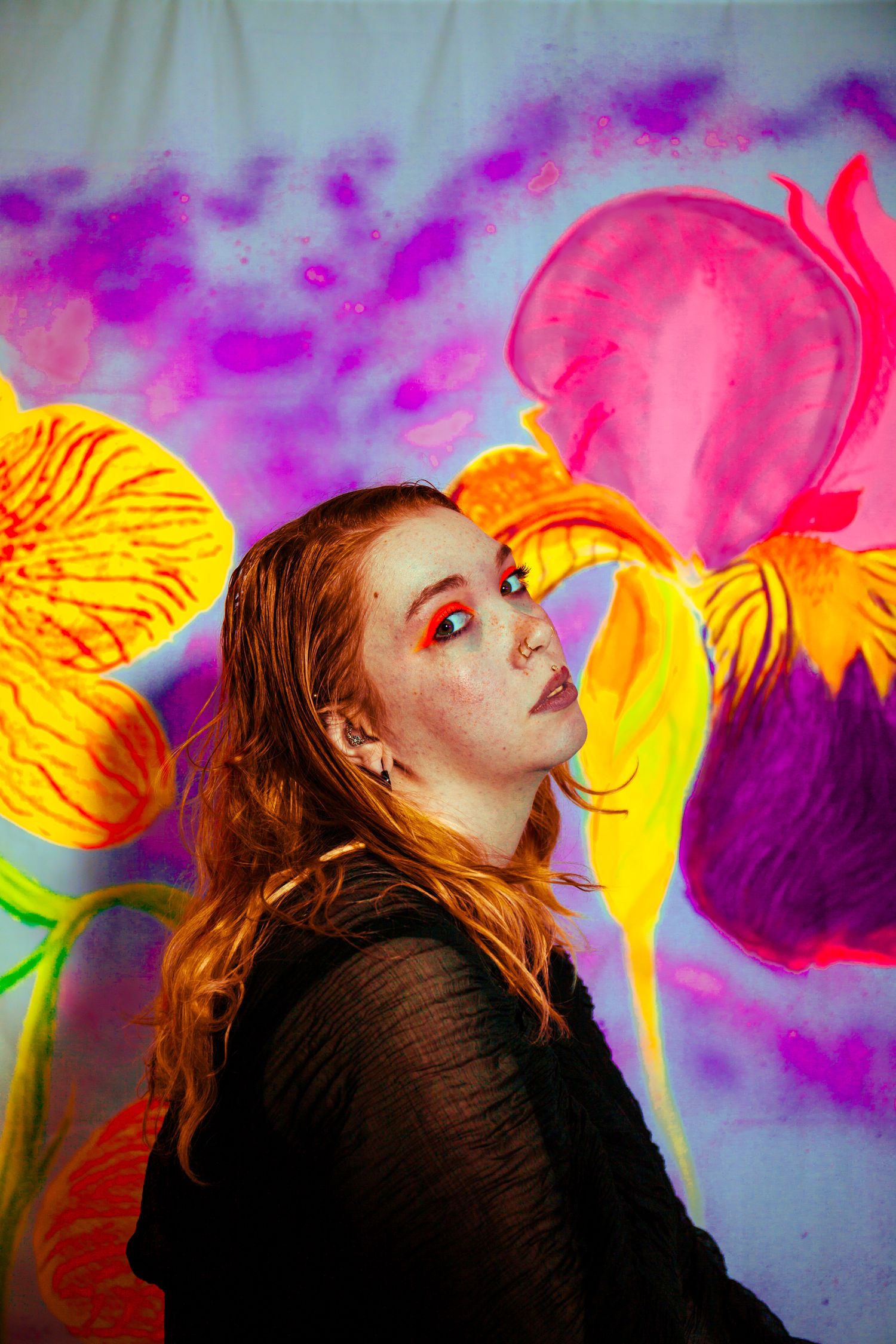 Redhead woman with bold orange eye makeup posing before vibrant floral painted backdrop.