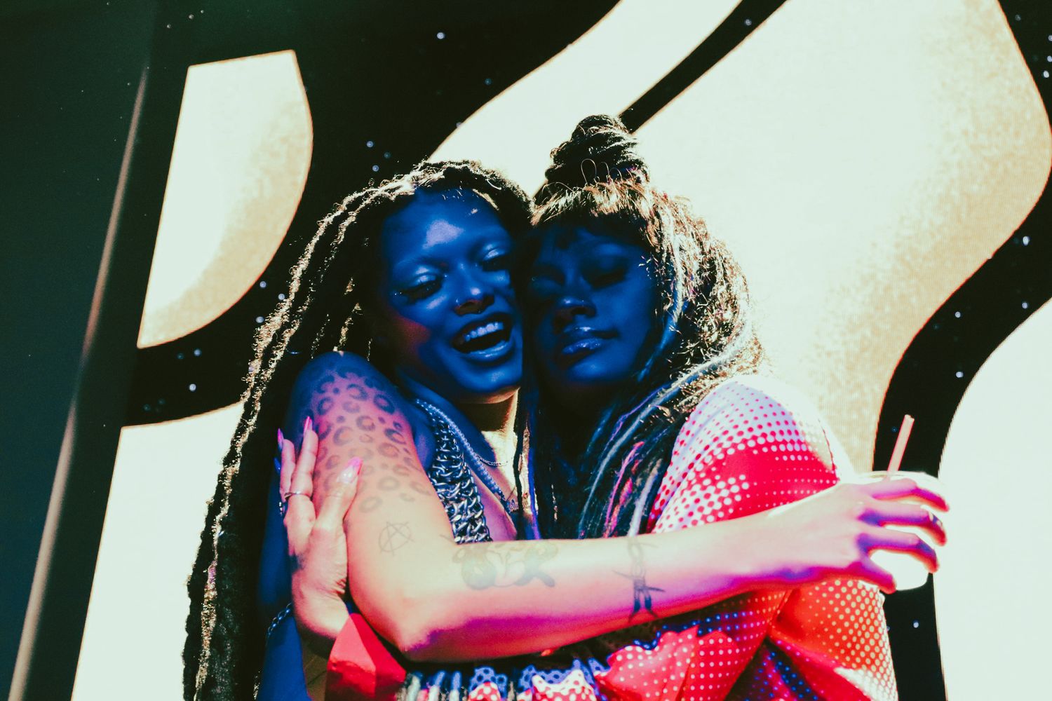 Two young Black women embrace and smile under dramatic blue stage lighting at an outdoor event.