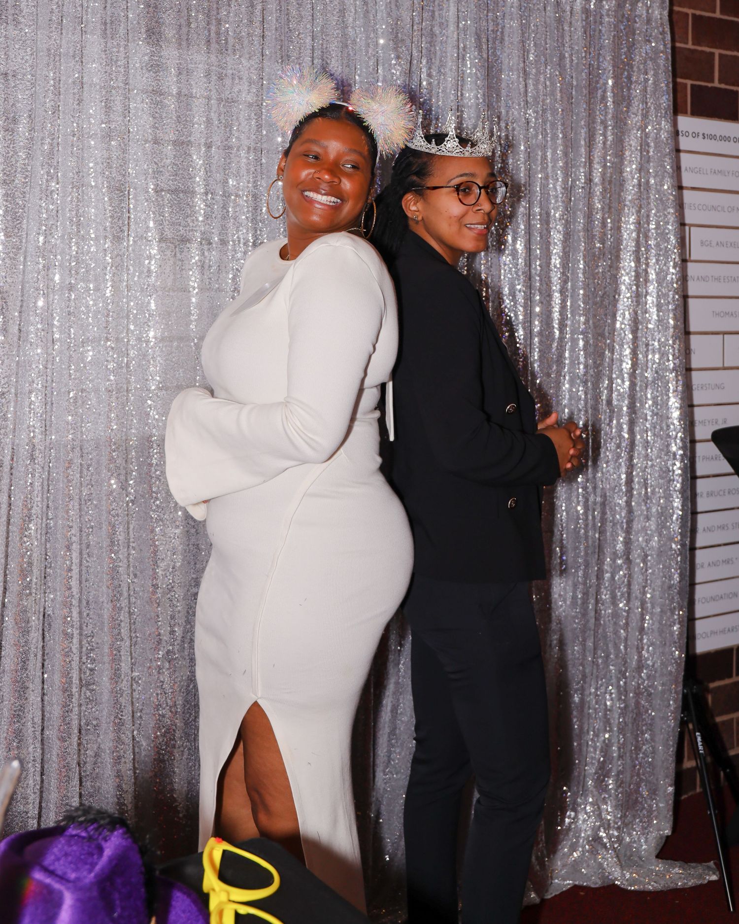 Two women posing back-to-back wearing crowns in front of silver sequin backdrop.