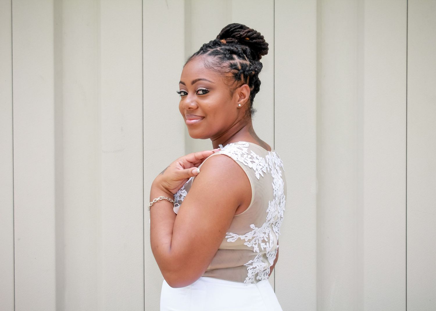 Elegant bride in white lace wedding gown posing with updo hairstyle against a neutral background.