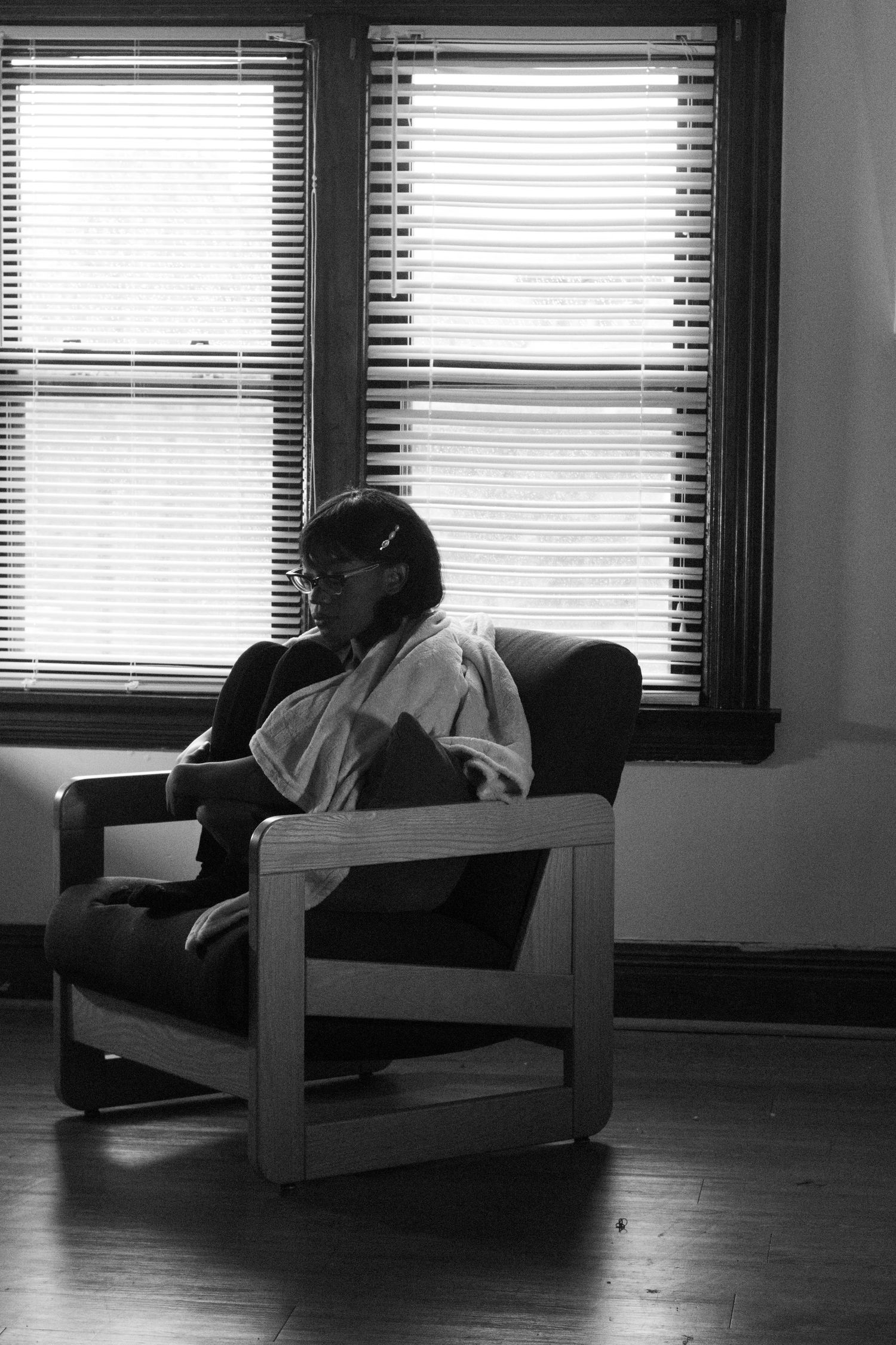 Black and white photo of a person sitting alone in an armchair reading near bright window blinds indoors.