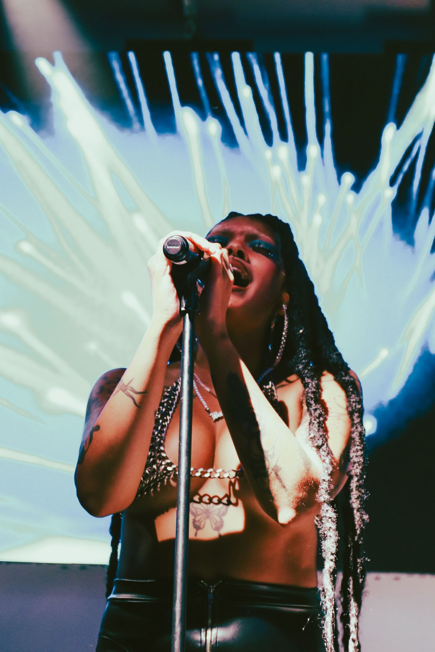 Female singer with locs performing on stage with microphone against vibrant light display backdrop.