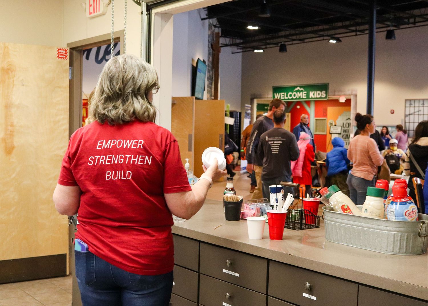 A volunteer in a red Empower Strengthen Build shirt stands at a counter in a community center.
