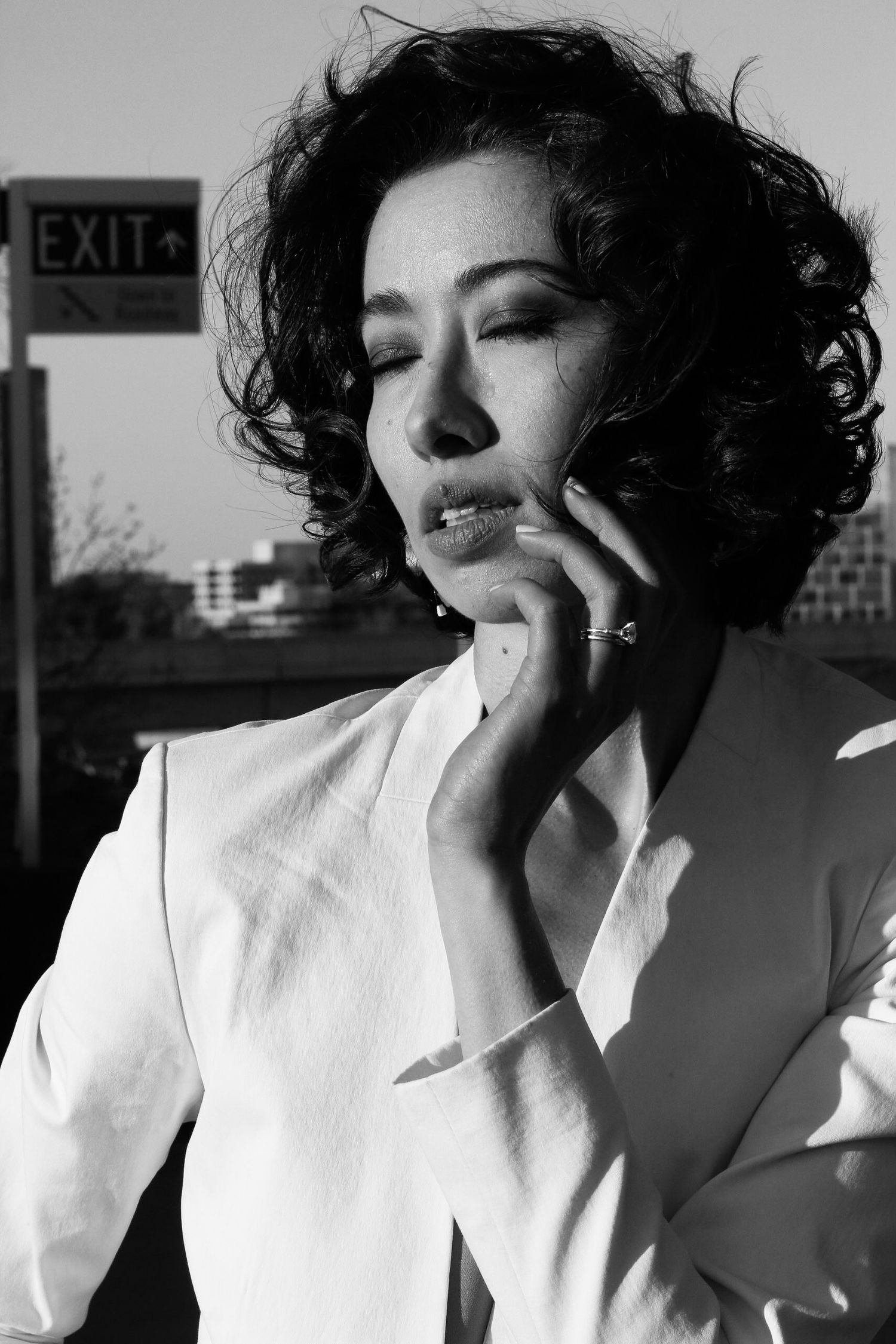 Black and white portrait of a curly-haired woman in a white blazer near an exit sign.