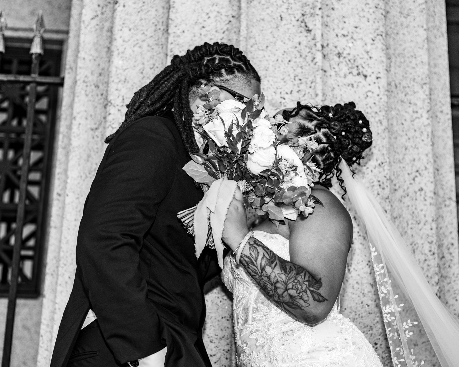 Black and white photo of a bride and groom sharing a kiss while holding a floral bouquet on their wedding day.
