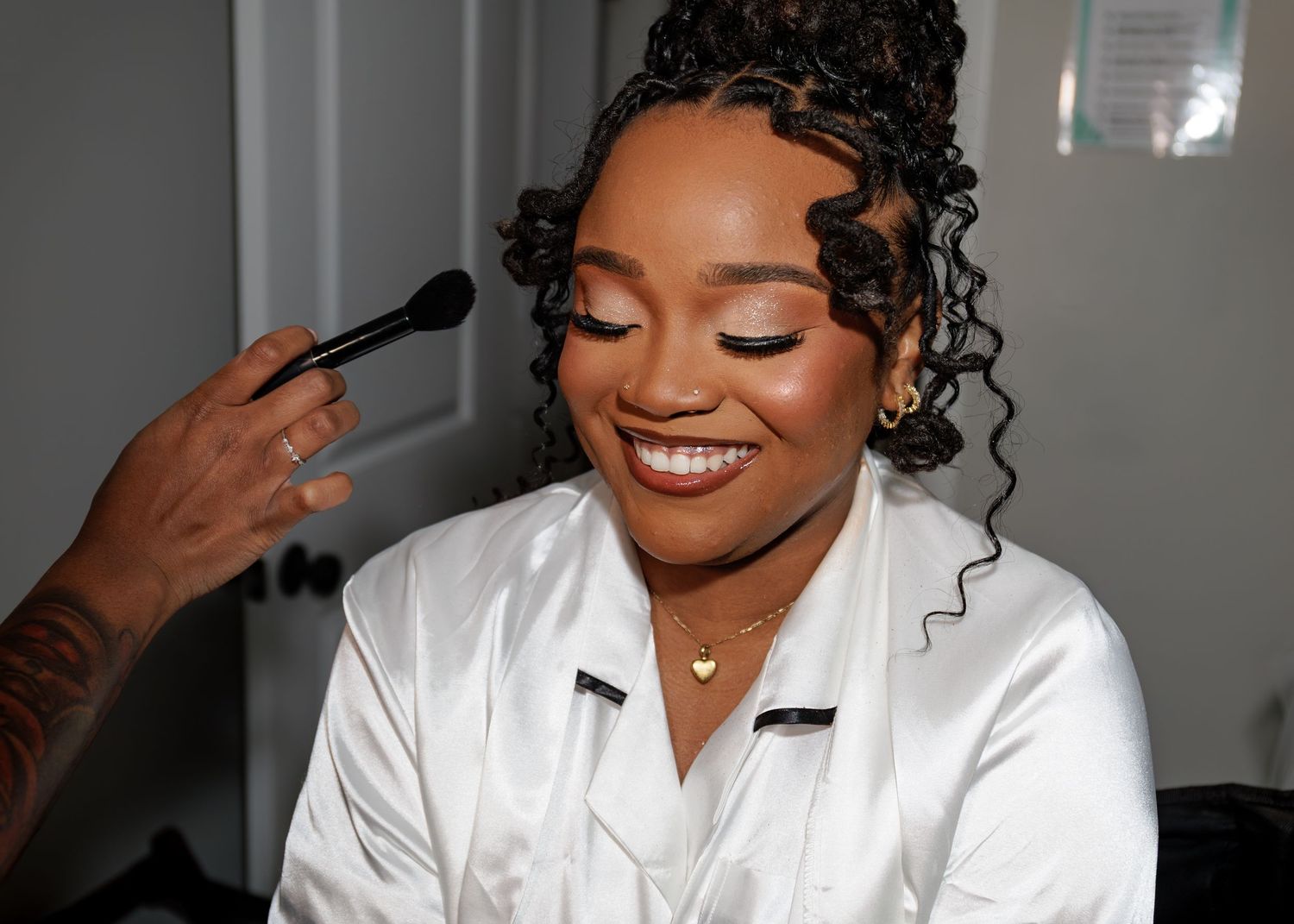 Smiling bride in a white robe gets makeup applied with a brush in a bright dressing room.