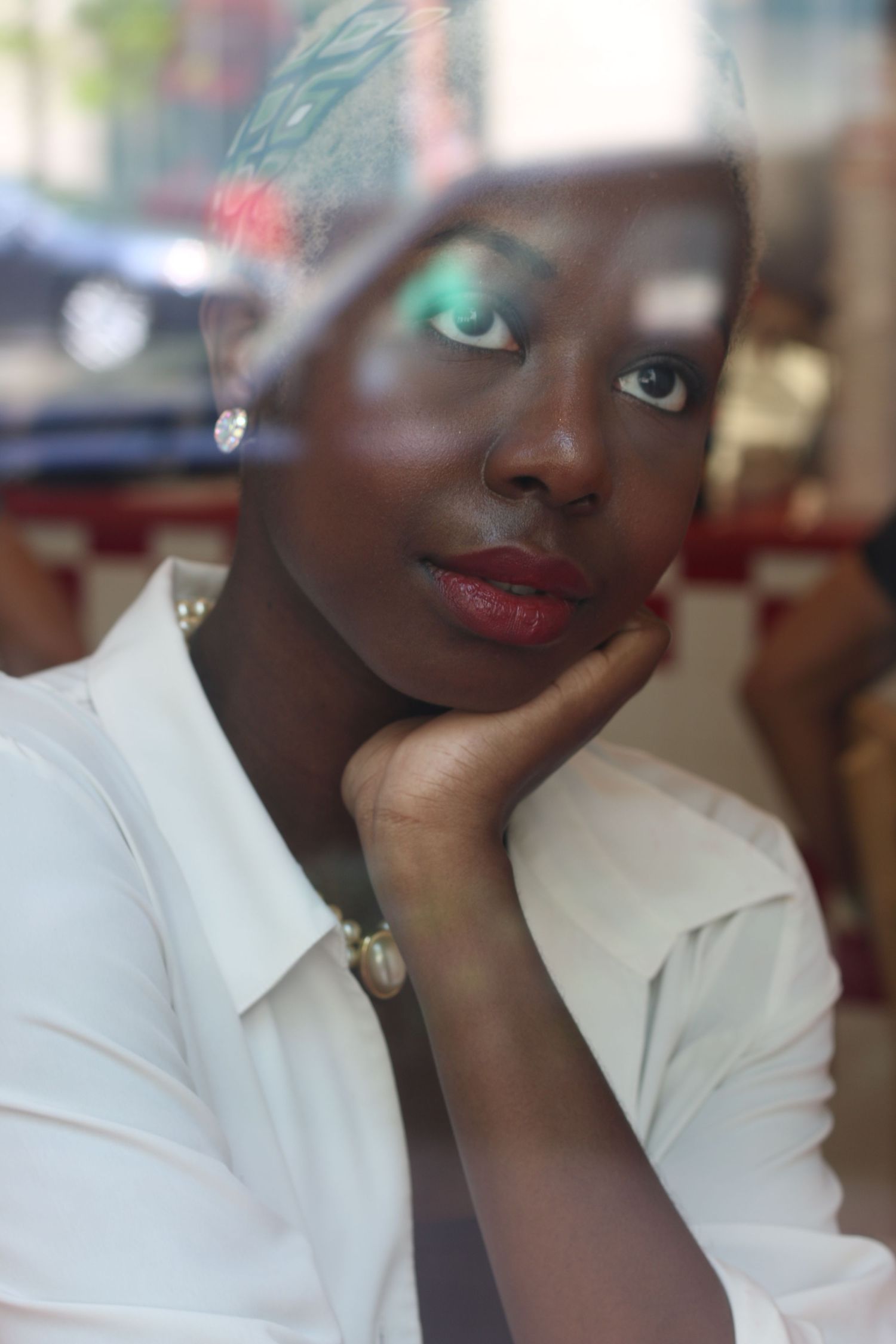 A dark-skinned Black woman in a white shirt with green eyeshadow and red lips poses behind a glass window.