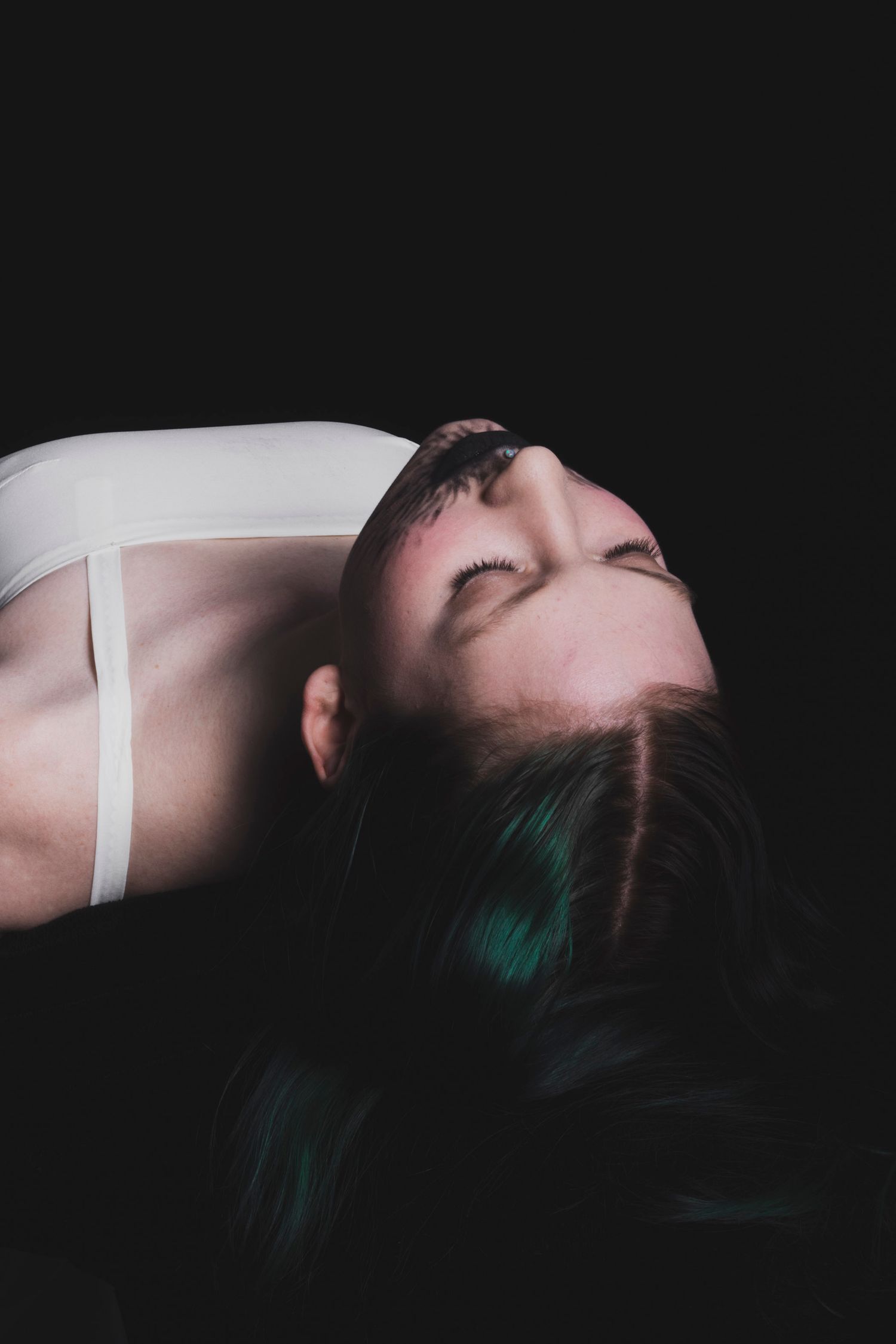 A woman with dark green hair lies back with eyes closed against a black background wearing a white top.