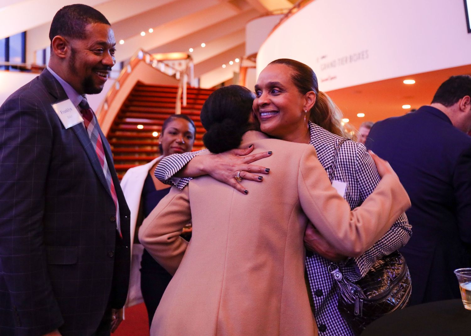 Woman in tan blazer hugging a guest at a professional networking reception.