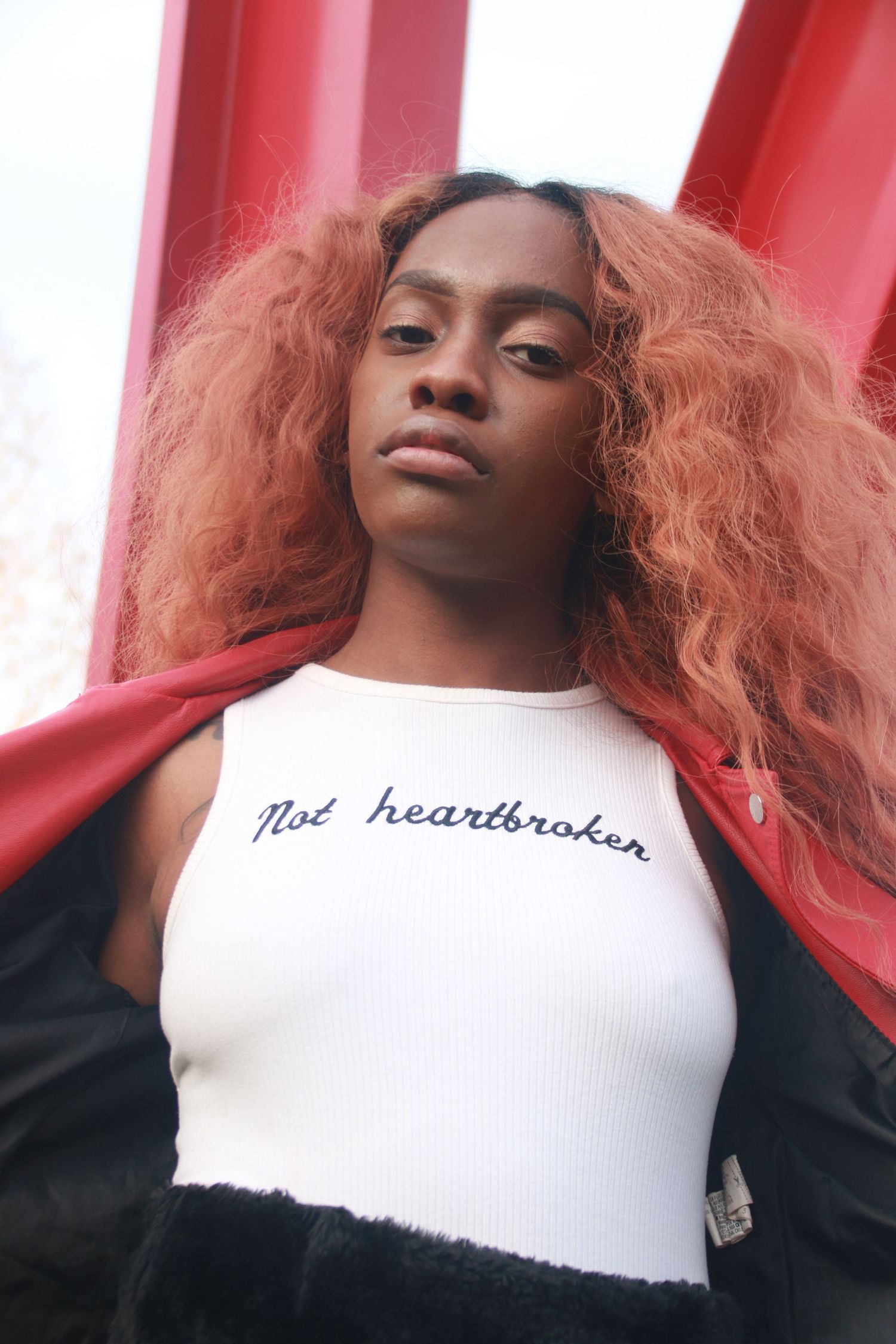 Young Black woman with red curly hair wears white 'Not Heartbroken' crop top near red structure.