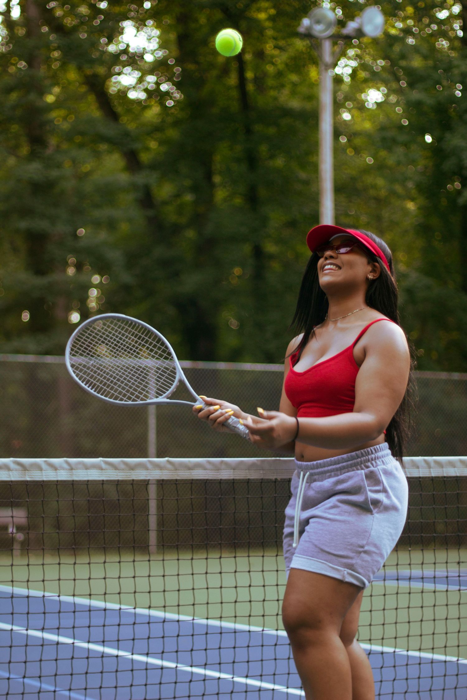 Woman in red crop top hitting tennis ball with racket on outdoor court near green trees.