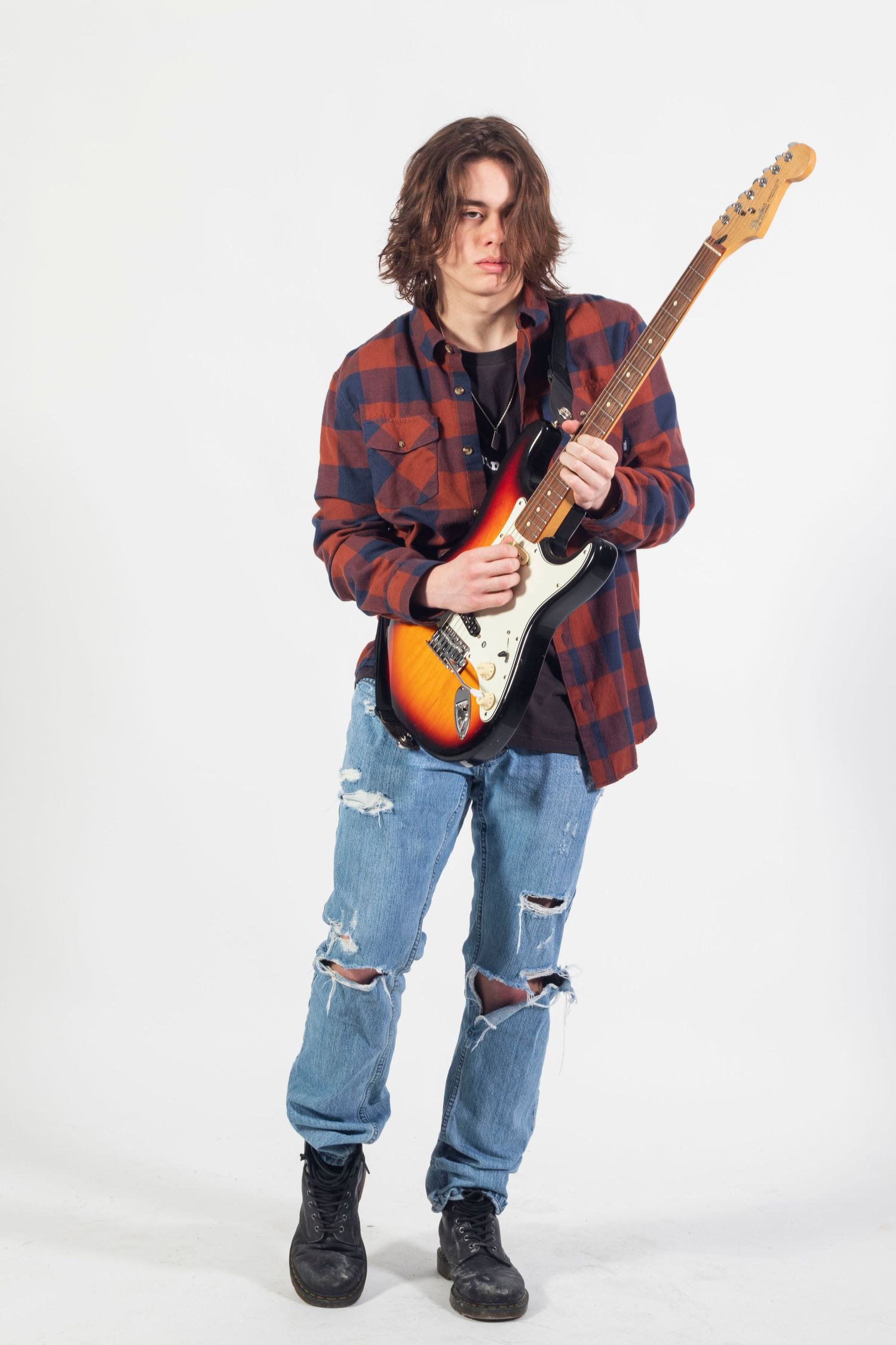 Young male guitarist with long hair plays a sunburst electric guitar wearing a plaid shirt and ripped jeans.