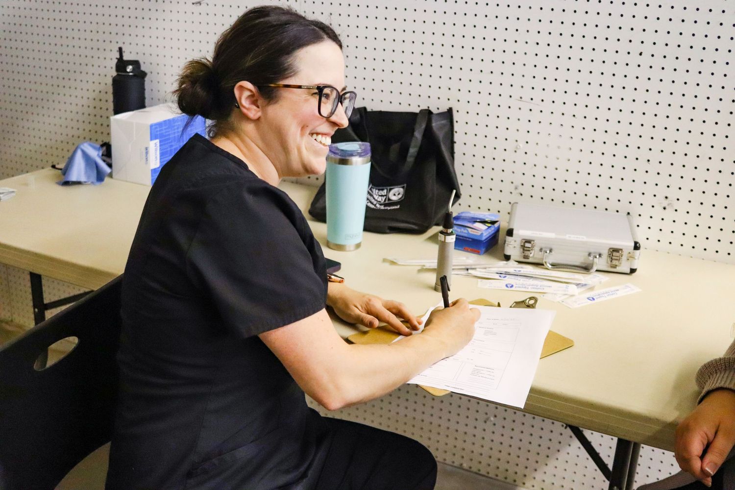 A smiling woman with glasses sits at a folding table reviewing paperwork in a workshop setting.