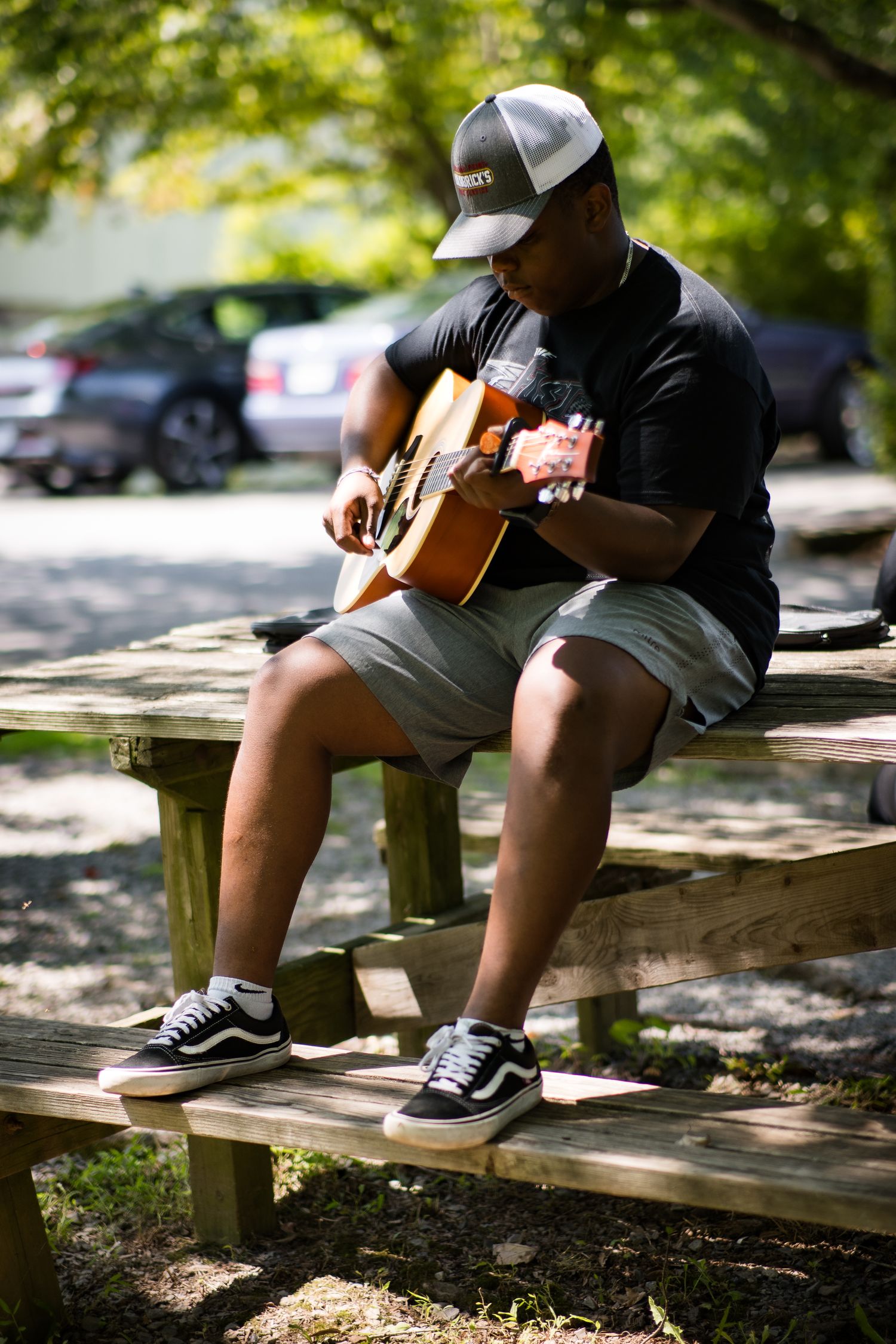 A young man sits on a wooden bench playing an acoustic guitar outdoors in a park.