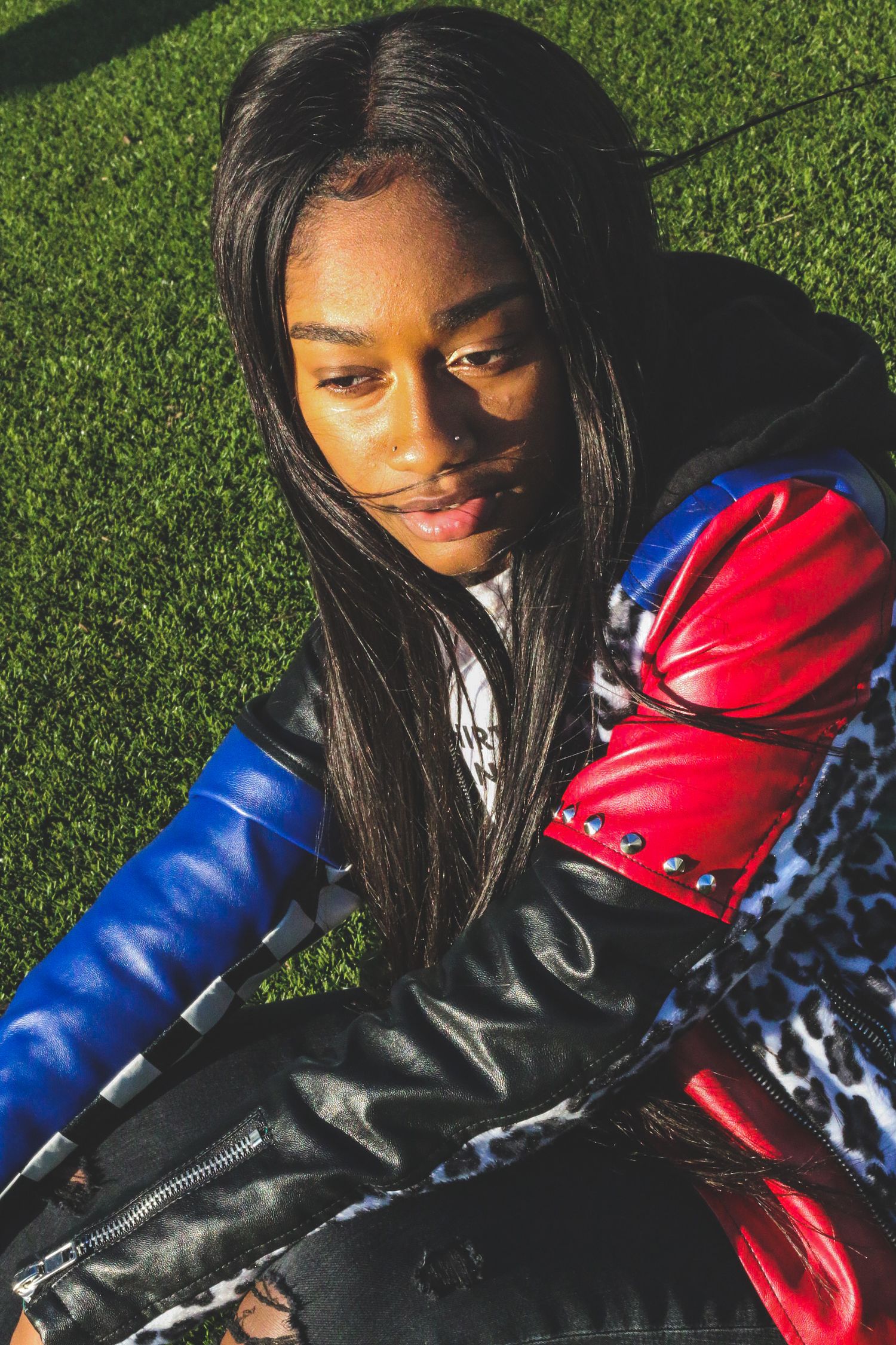 Young Black woman in colorful leather jacket sitting on green grass.