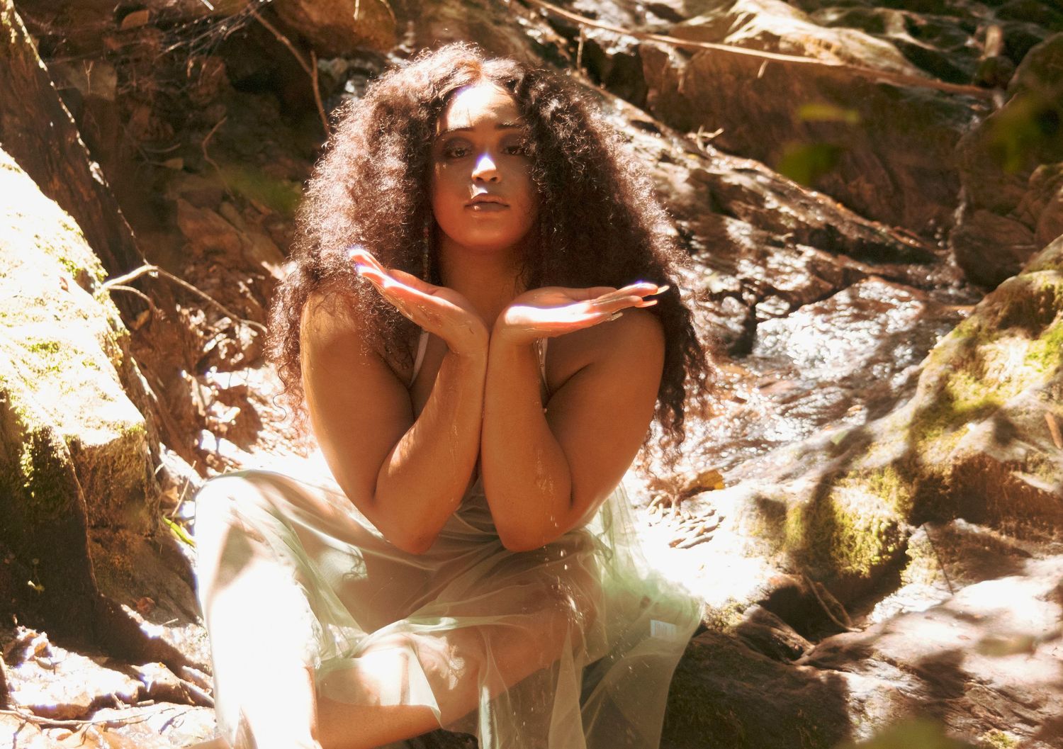 Woman with curly hair poses gracefully among rocks and moss in a dreamy outdoor setting.