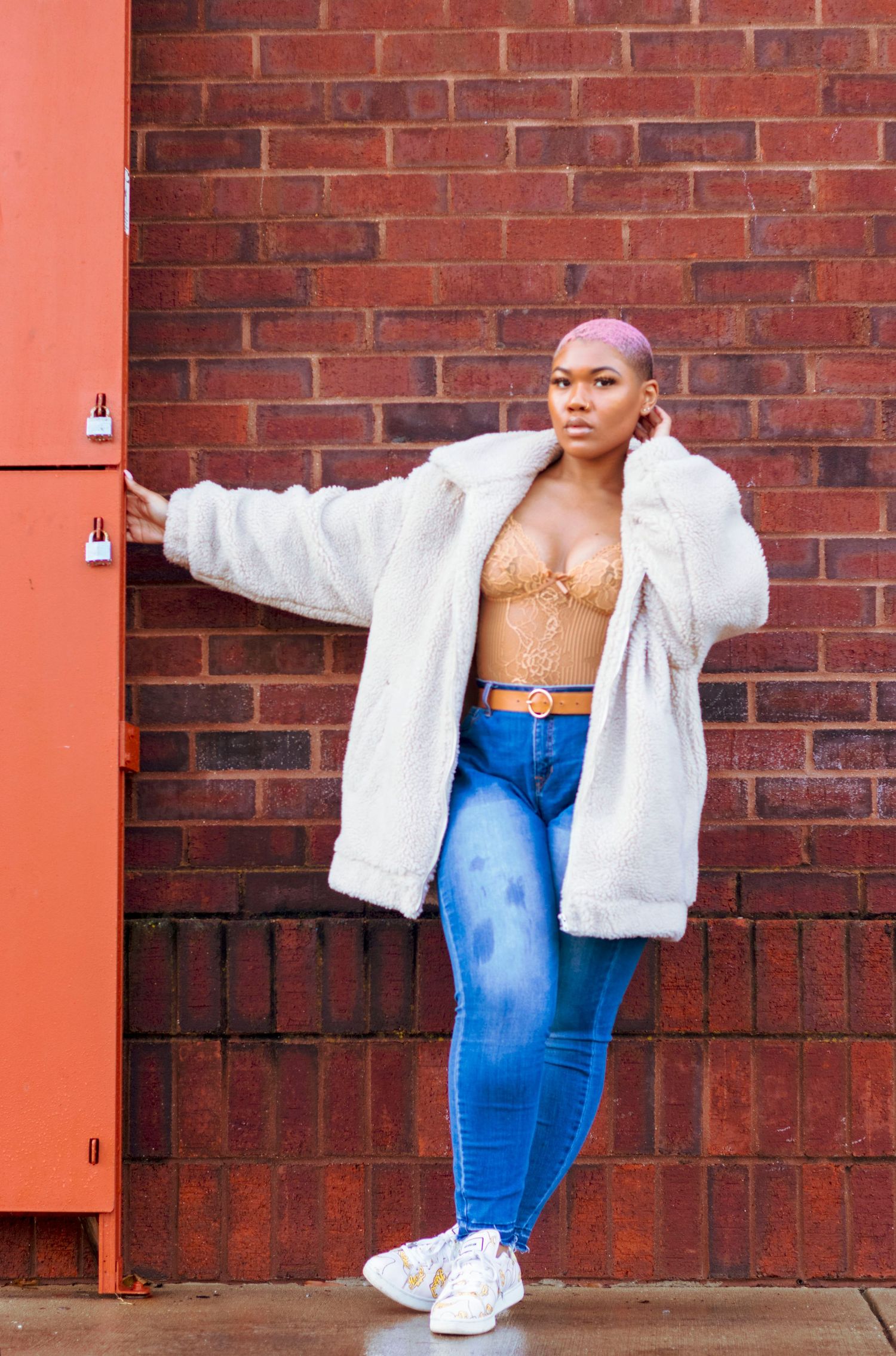 Confident Black woman with pink hair in white faux fur coat and jeans posing against red brick wall.