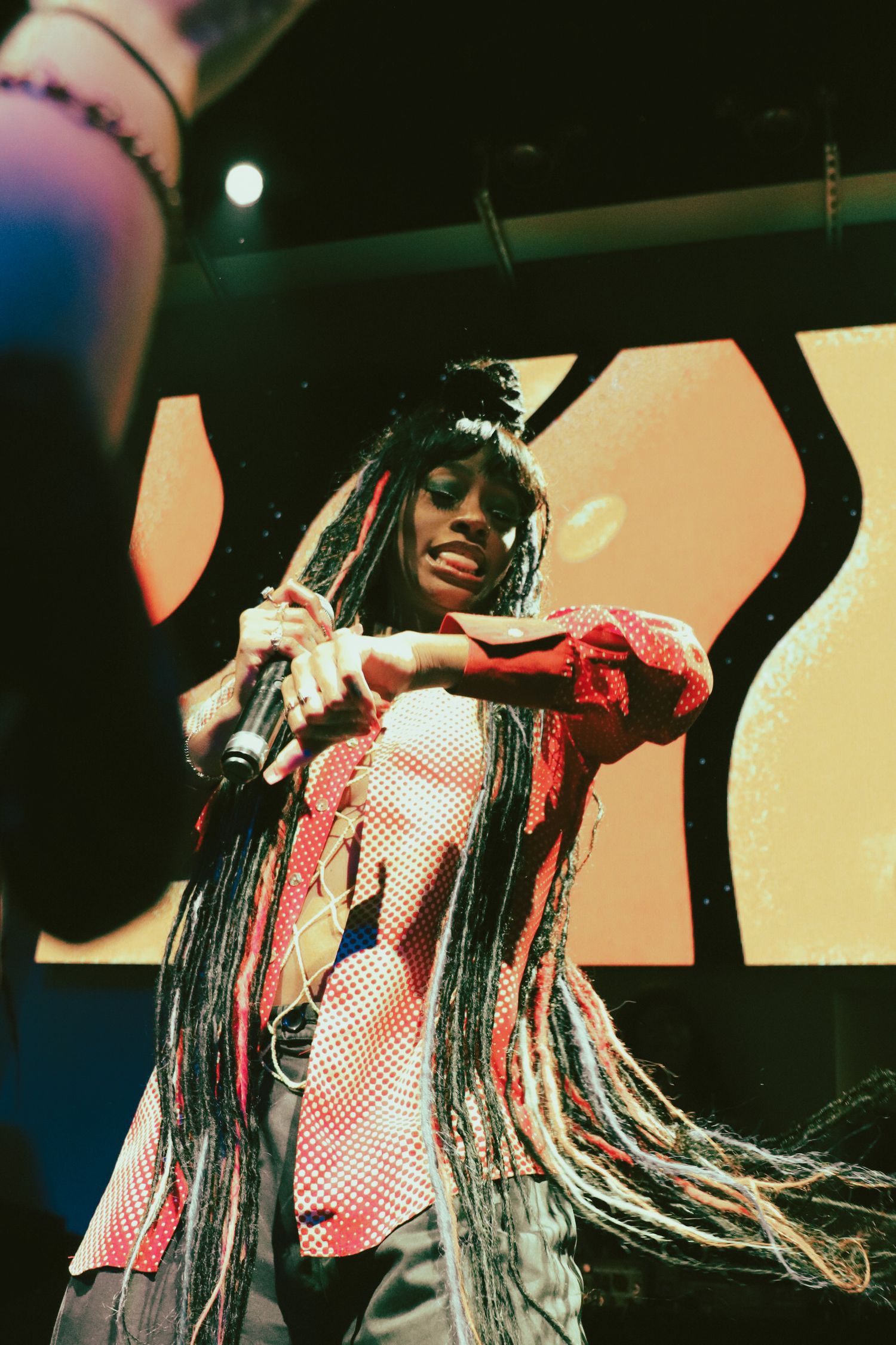 Female performer with long multicolored braids singing passionately on stage at a live concert.