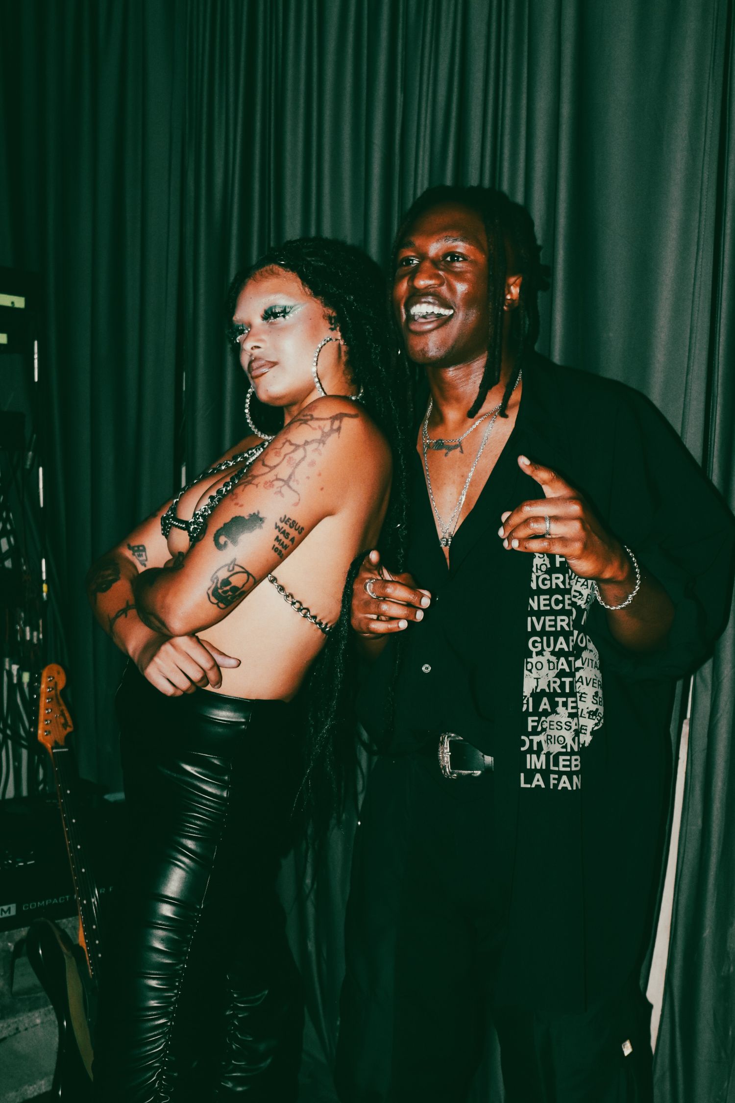 Two performers posing backstage at a concert, woman with tattoos in leather and man in printed shirt.