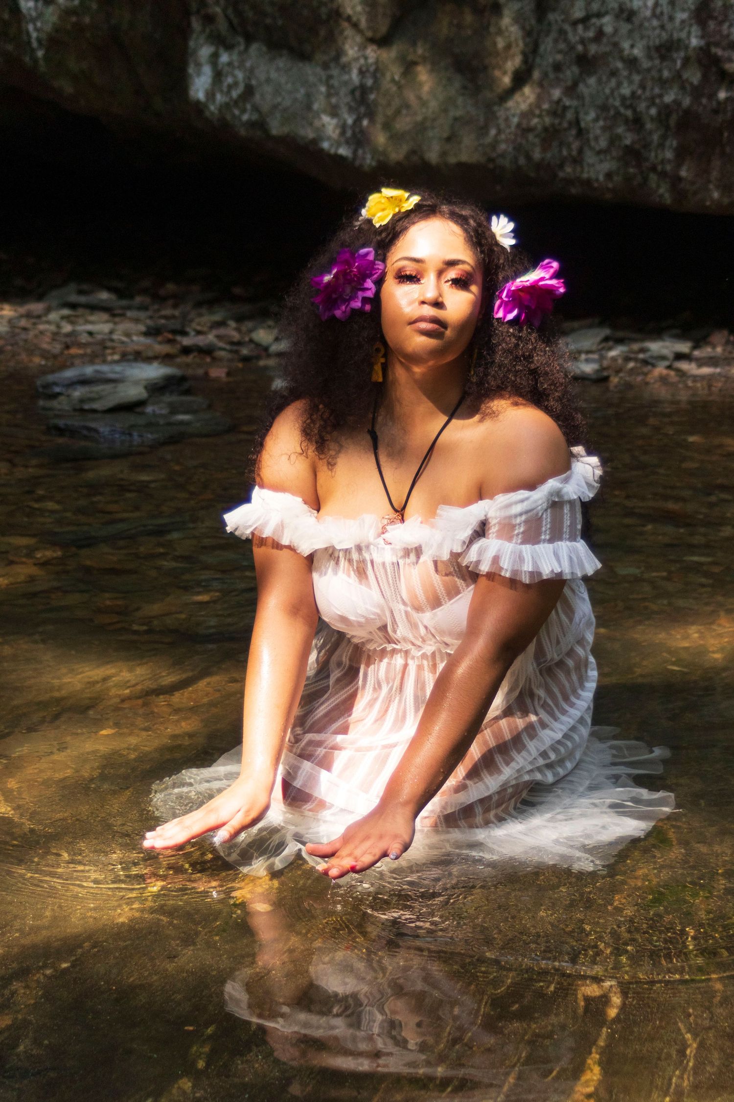A woman with flowers in her curly hair kneels in shallow water wearing a sheer white off-shoulder dress.
