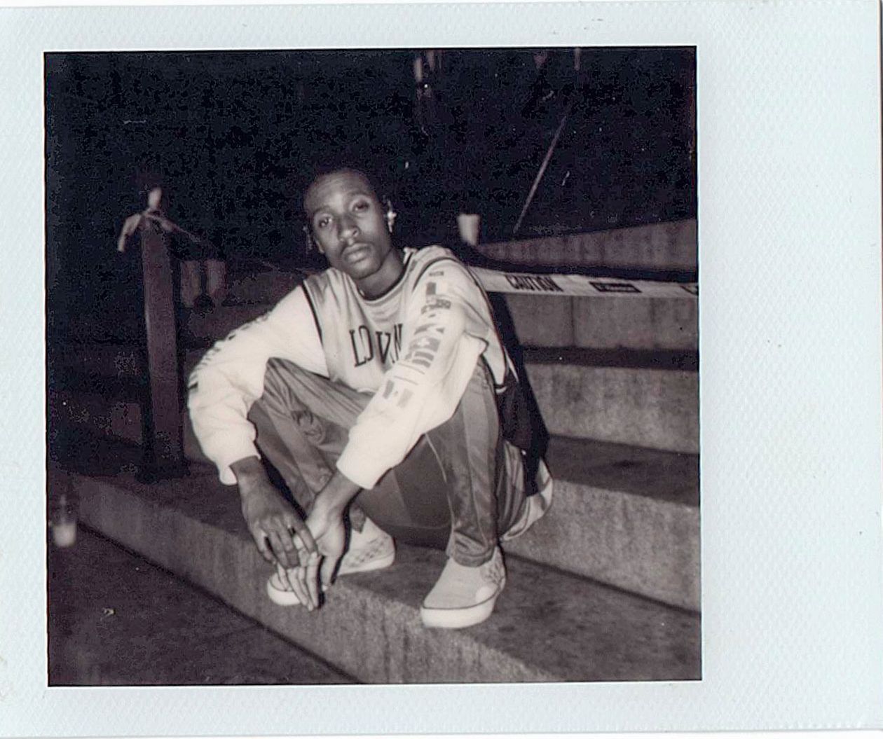 Young man crouching on outdoor steps at night in a white sweatshirt and jeans in a black and white polaroid photo.