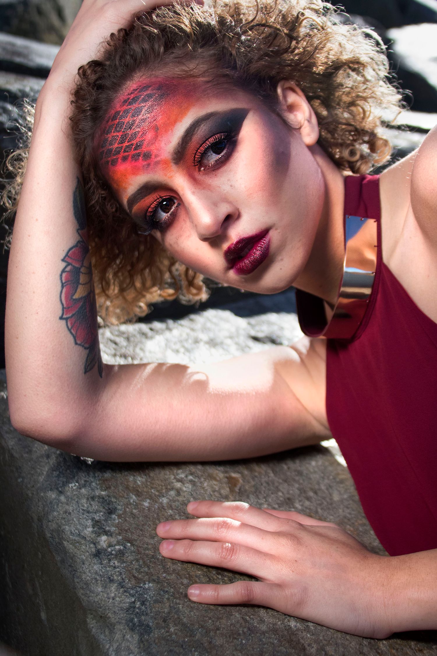 A woman with curly hair showcases dramatic red and black scale-patterned avant-garde makeup against rocky terrain.