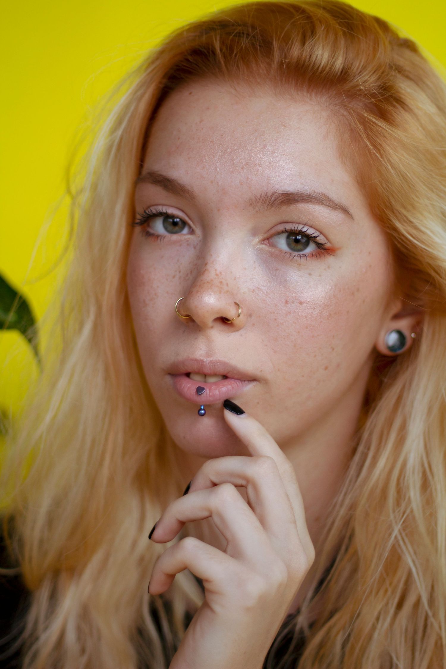 Young blonde woman with freckles and facial piercings posing against a yellow background.