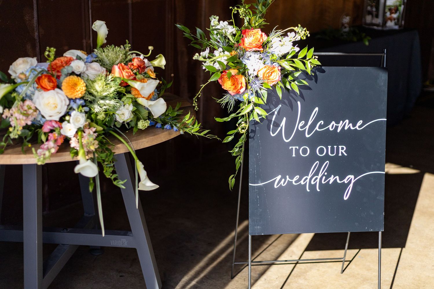 Elegant wedding welcome sign on easel beside colorful floral arrangement at venue entrance.