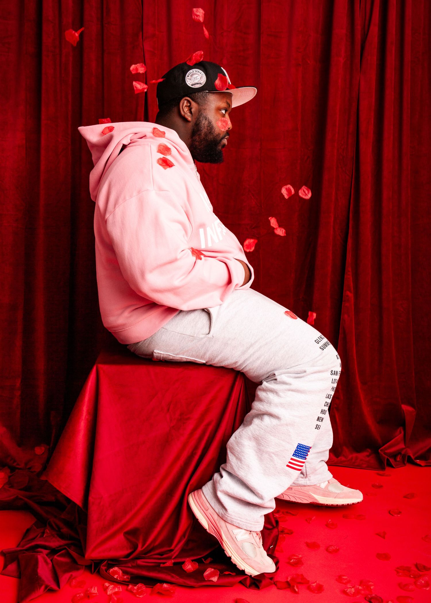 Bearded Black man in pink hoodie sits on red draped cube with falling rose petals backdrop.
