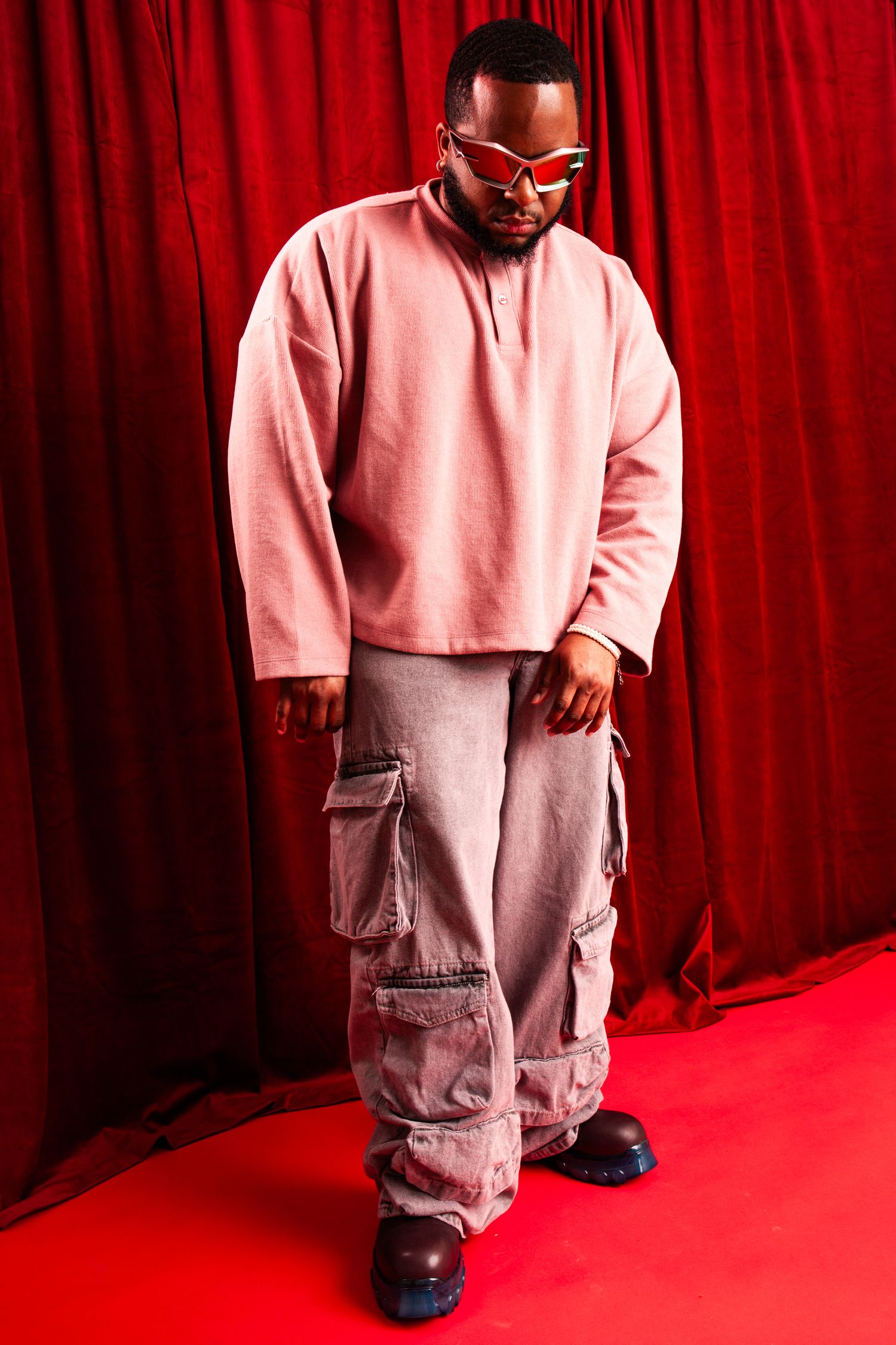 Stylish man in pink oversized sweatshirt and cargo pants with futuristic sunglasses on red backdrop.