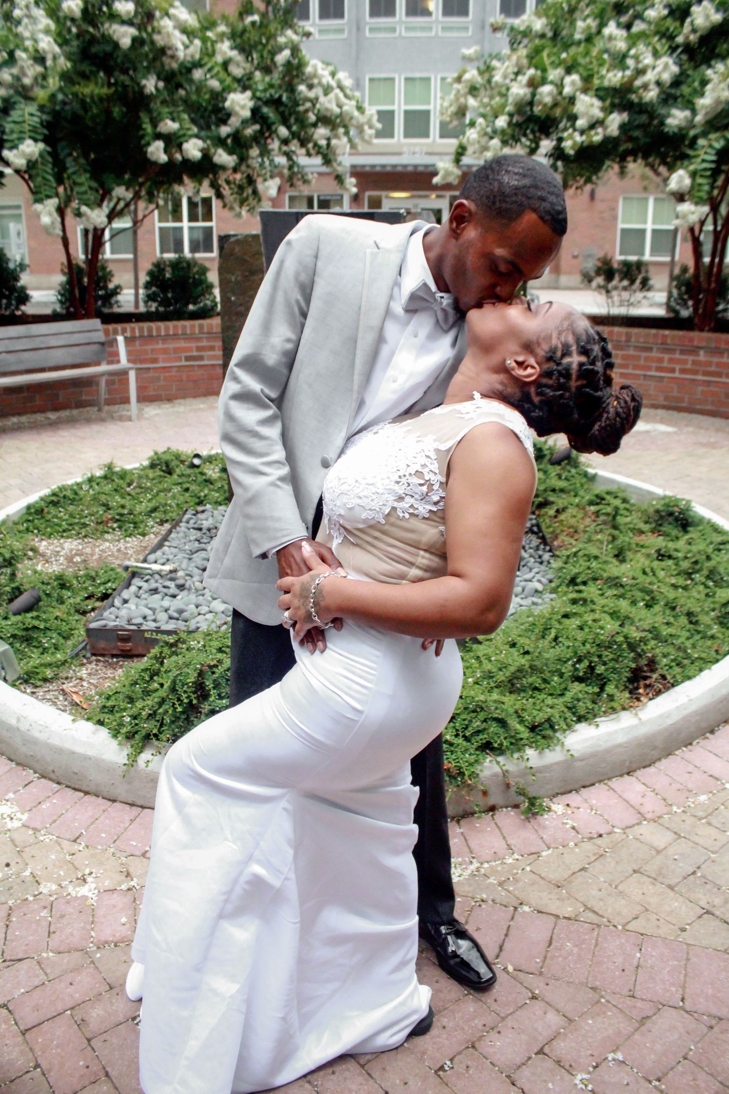 Bride and groom sharing romantic kiss outdoors in courtyard with blooming white flowers in background.