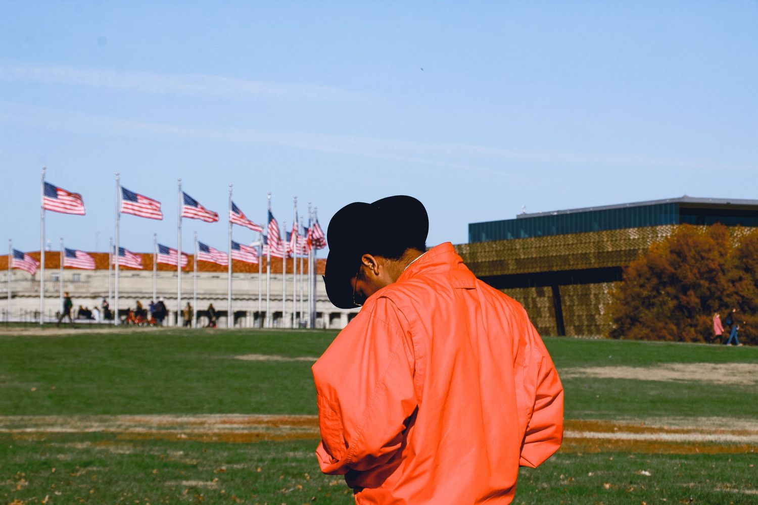 Person in orange jacket stands before American flags at outdoor monument on sunny day.