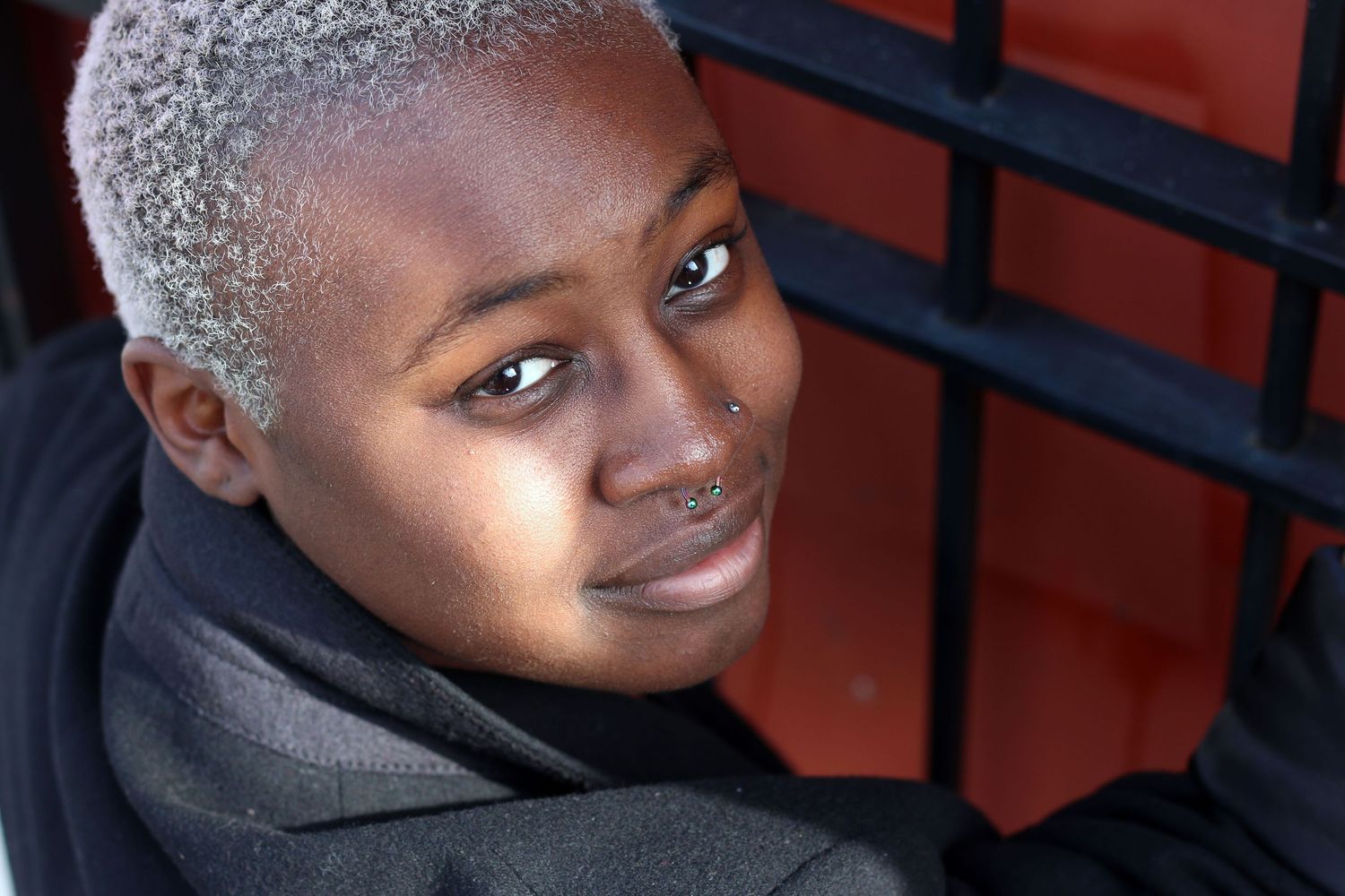 A young Black person with short silver hair and a nose ring leans against a red metal structure.