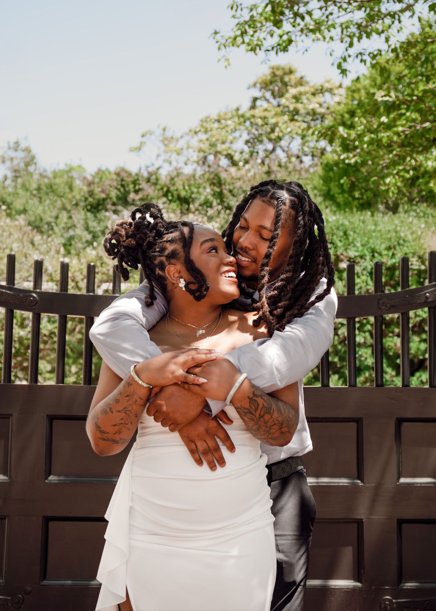 A joyful couple in wedding attire embraces outdoors, the groom hugging the bride from behind near a dark fence.