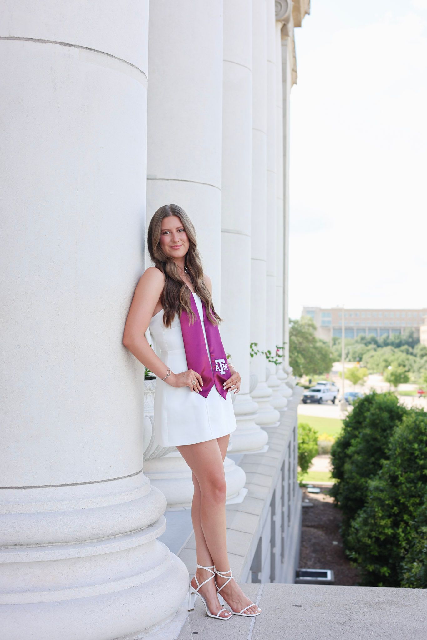 Young woman in white dress and maroon graduation stole poses between white columns at Texas A&M University.