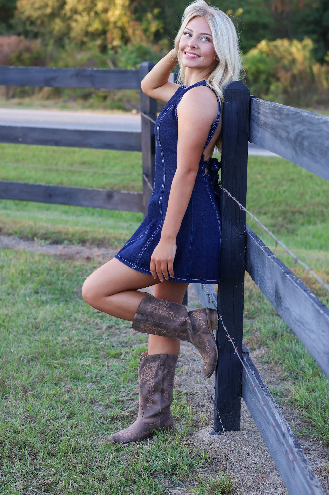 Blonde woman in blue denim dress and brown cowboy boots leaning against a rustic wooden fence.