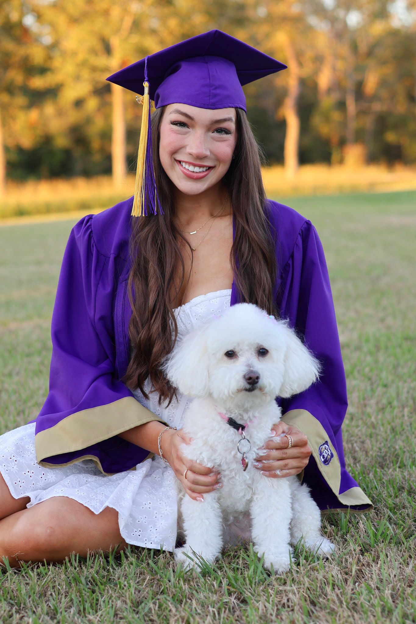 Smiling graduate in purple cap and gown sitting on grass holding a white fluffy dog outdoors.