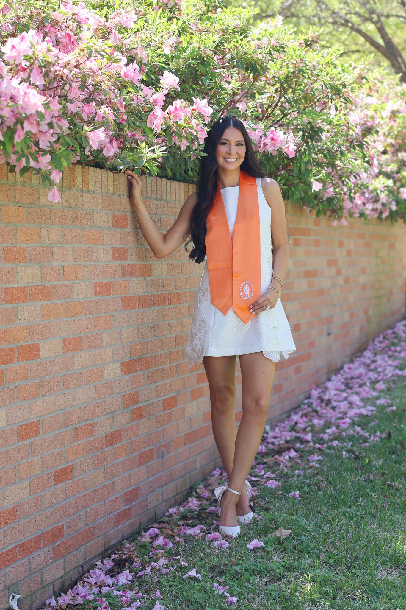 Young woman in white dress and orange graduation stole poses by brick wall with blooming pink flowers.