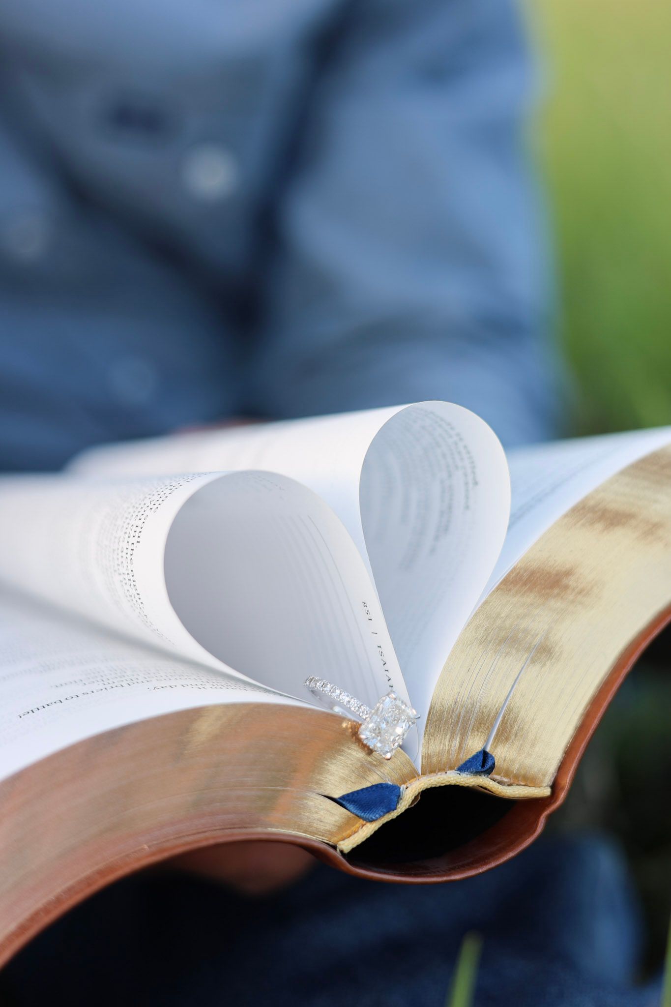 Bible pages folded into a heart shape with wedding rings resting in the center, held by a person outdoors.
