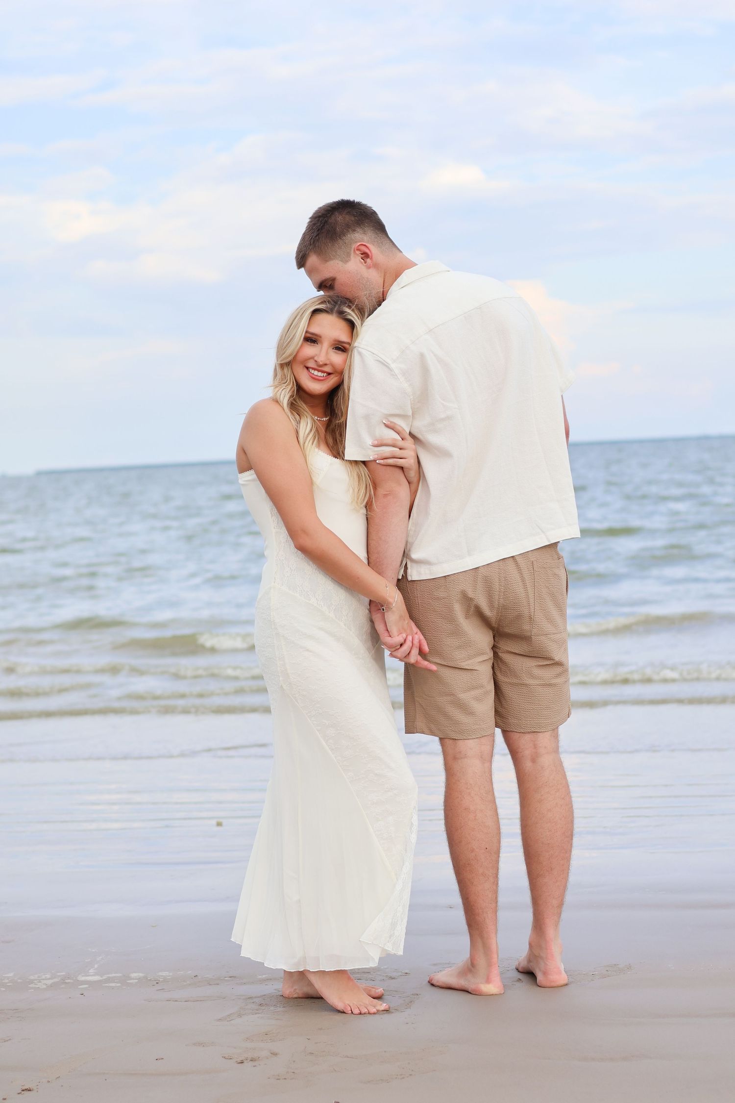 A couple embraces on a beach at sunset, with the woman wearing a white dress and the man in khaki shorts.